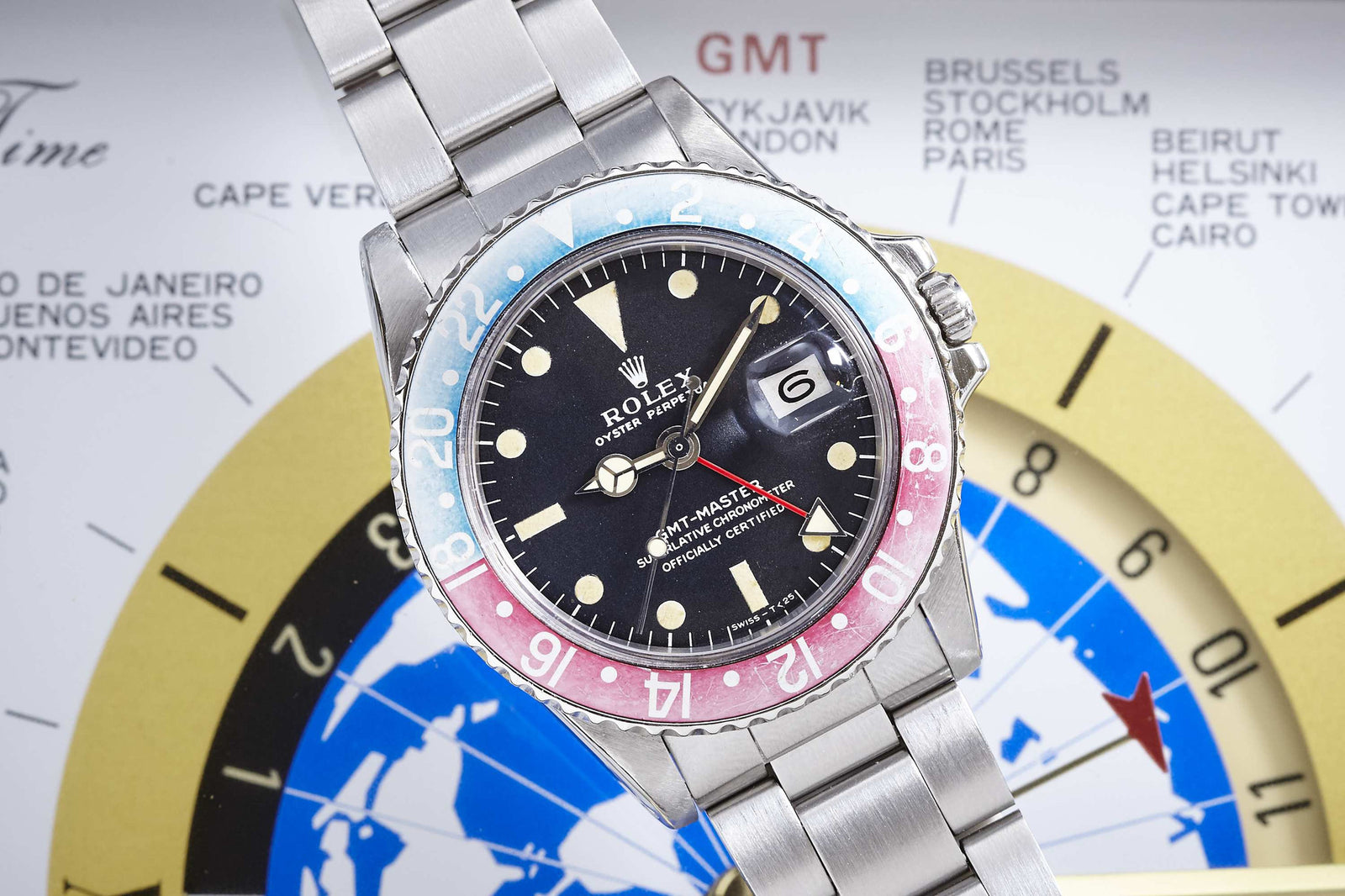 A close-up of the Rolex GMT Master 1675 daily wear watch by Rolex, featuring a red and blue bezel, black dial, and stainless steel band, set against a world time map displaying city names and time zones.