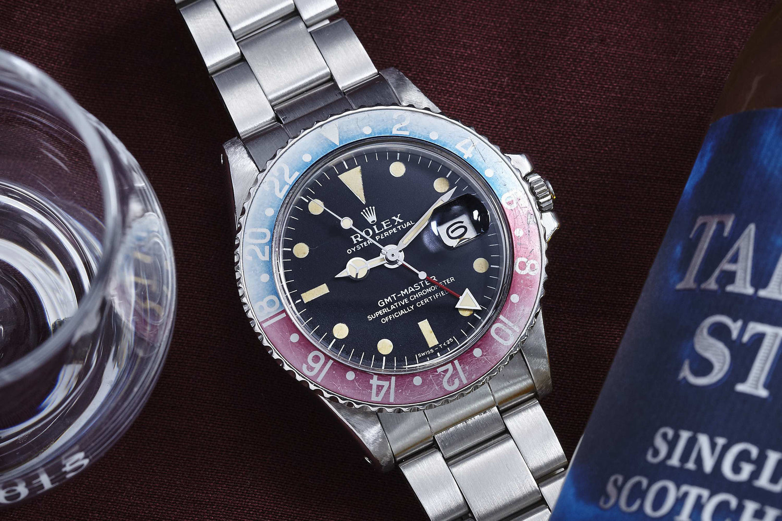 The Rolex GMT Master 1675 by Rolex, featuring a red and blue bezel with a silver metal band, sits on dark fabric beside a glass and Scotch whisky—a timeless daily-wear watch perfect for vintage enthusiasts.