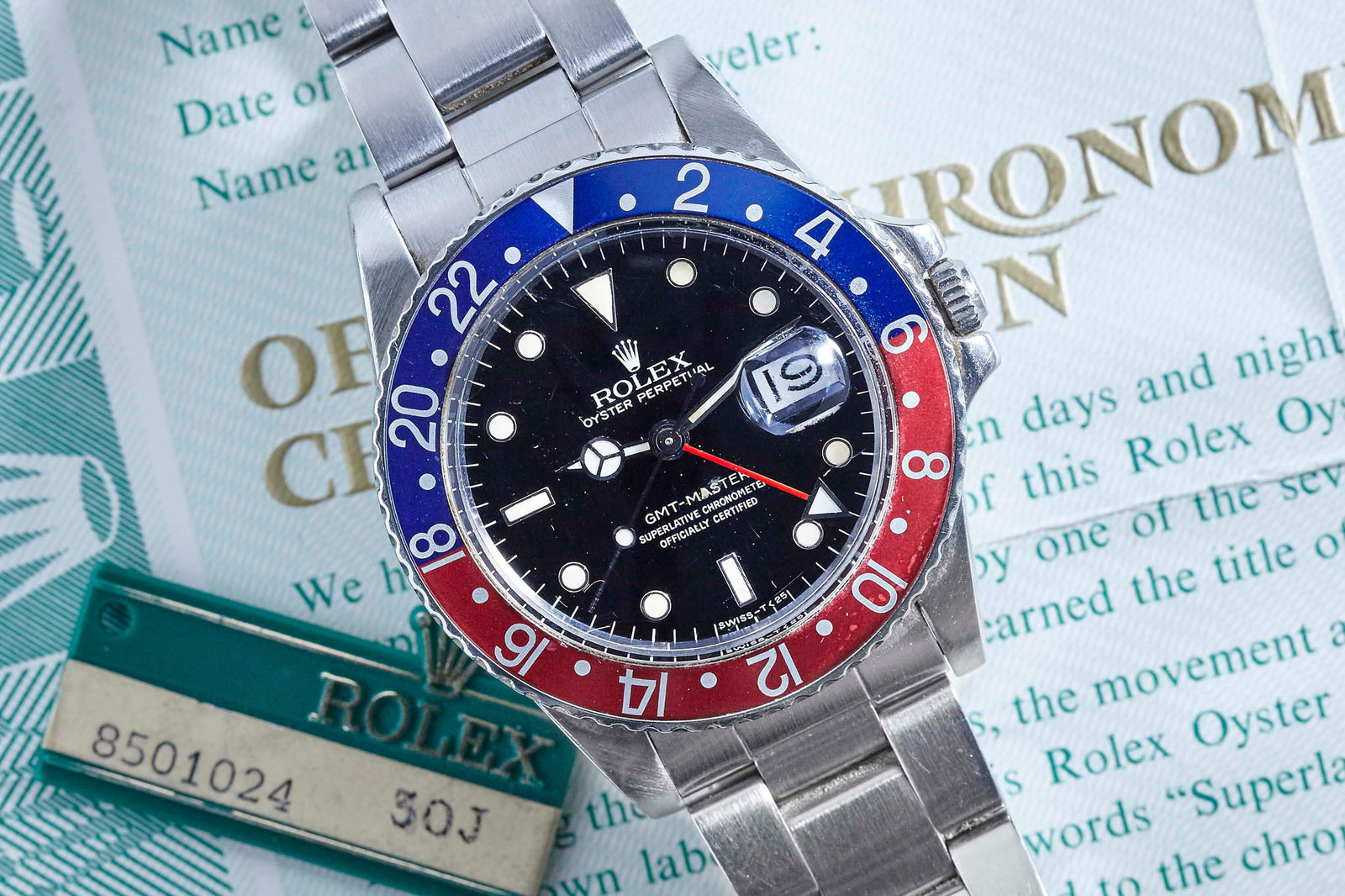 A Rolex GMT Master 16750 with red and blue bezel rests atop its certification papers and a green Rolex tag bearing serial no. 8501024. The black dial features a date display. Detailed description coming soon!.