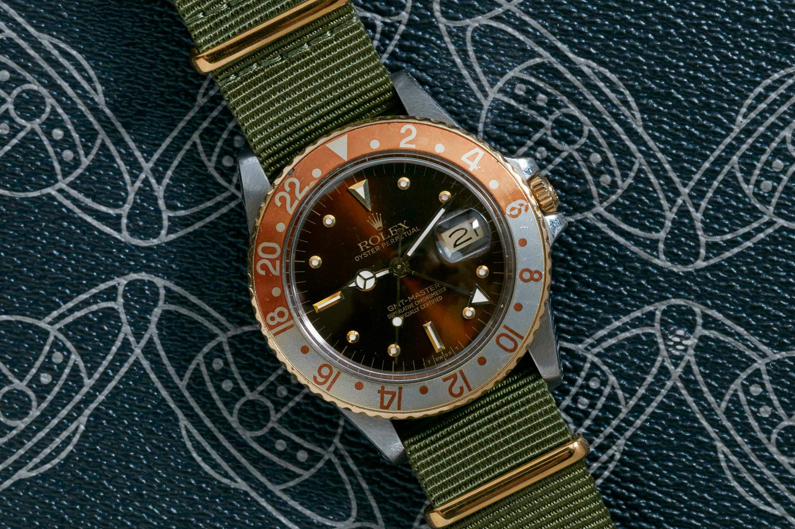The Rolex GMT Master 'Rootbeer' by Rolex, featuring a stainless steel case, brown and gold bezel, and green fabric strap, is displayed on a textured surface with the watch face showing the date and time.