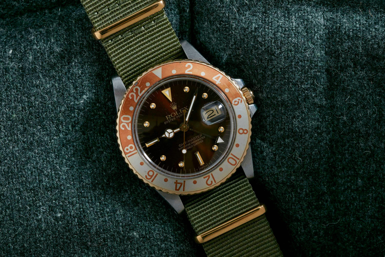 A Rolex GMT Master 'Rootbeer' watch by Rolex, featuring a brown and gold bezel, black dial with gold accents, and green nylon strap, is displayed on textured dark green fabric. The date shows on the stainless steel case.