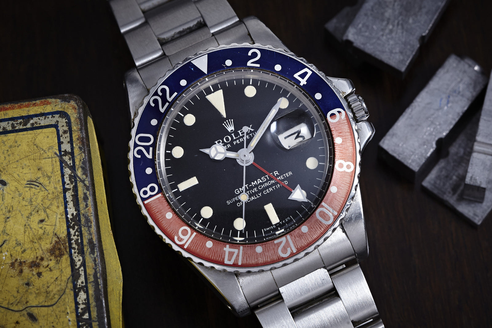 The Rolex GMT Master 1675 -1969, a classic vintage timepiece from Rolex, with its stainless steel band and red-blue bezel, sits on a dark surface beside metal parts and a worn yellow box. The watch displays the time at approximately 2:08.