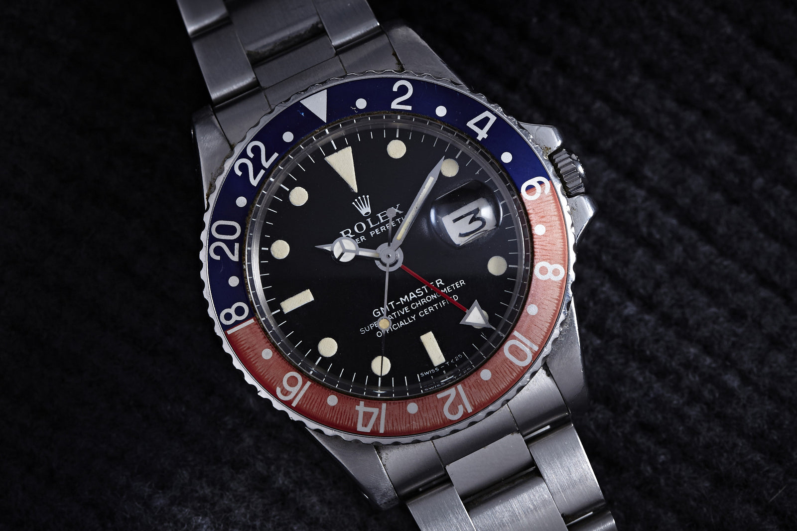 A close-up of the Rolex GMT Master 1675 -1969, featuring a stainless steel band, black dial, and red-blue bezel set to 10:10 and date 10, resting on a dark textured surface.