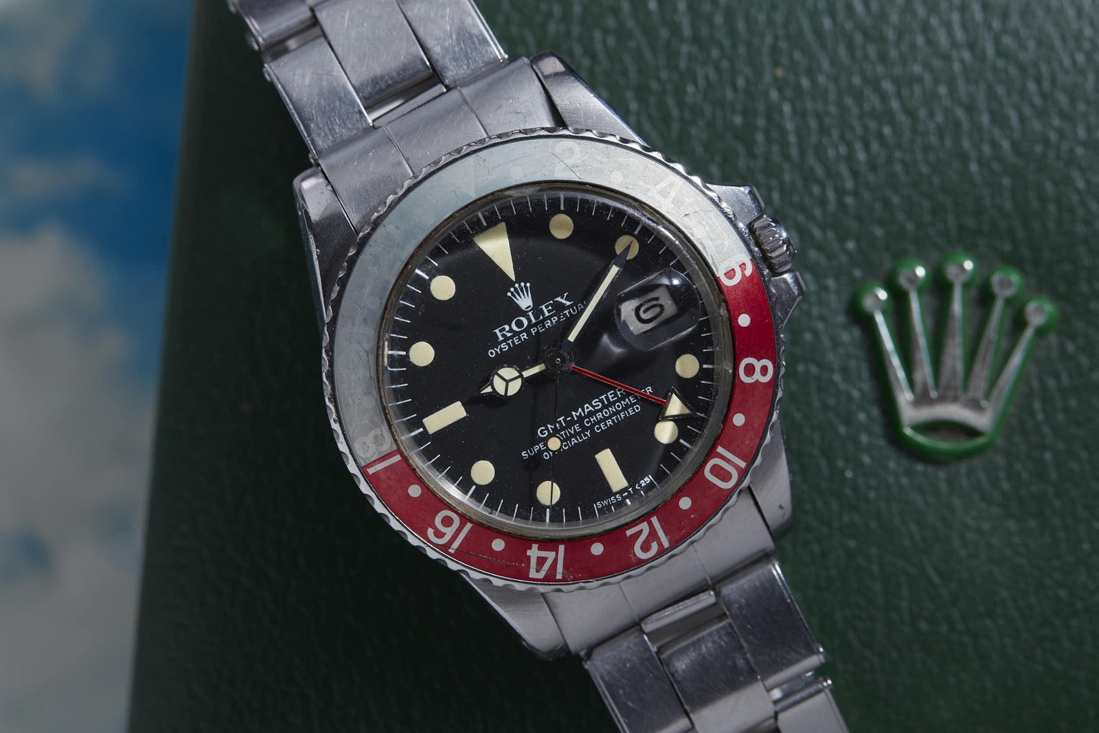 A Rolex GMT Master watch by Rolex featuring a red and black bezel, black dial, and metal band is displayed on a green surface with the silver Rolex crown logo nearby.