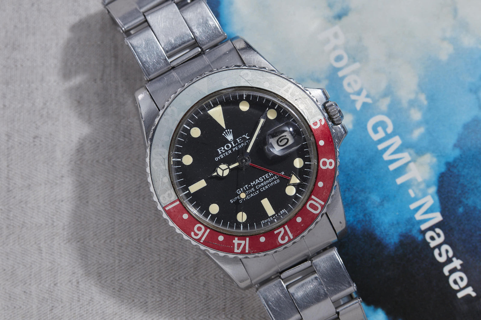 A Rolex GMT Master wristwatch with a red and black bezel and metal strap rests on its branded booklet with a cloudy sky, evoking the watch's transcontinental travel heritage; the date reads 6. Brand: Rolex. Product: Rolex GMT Master.