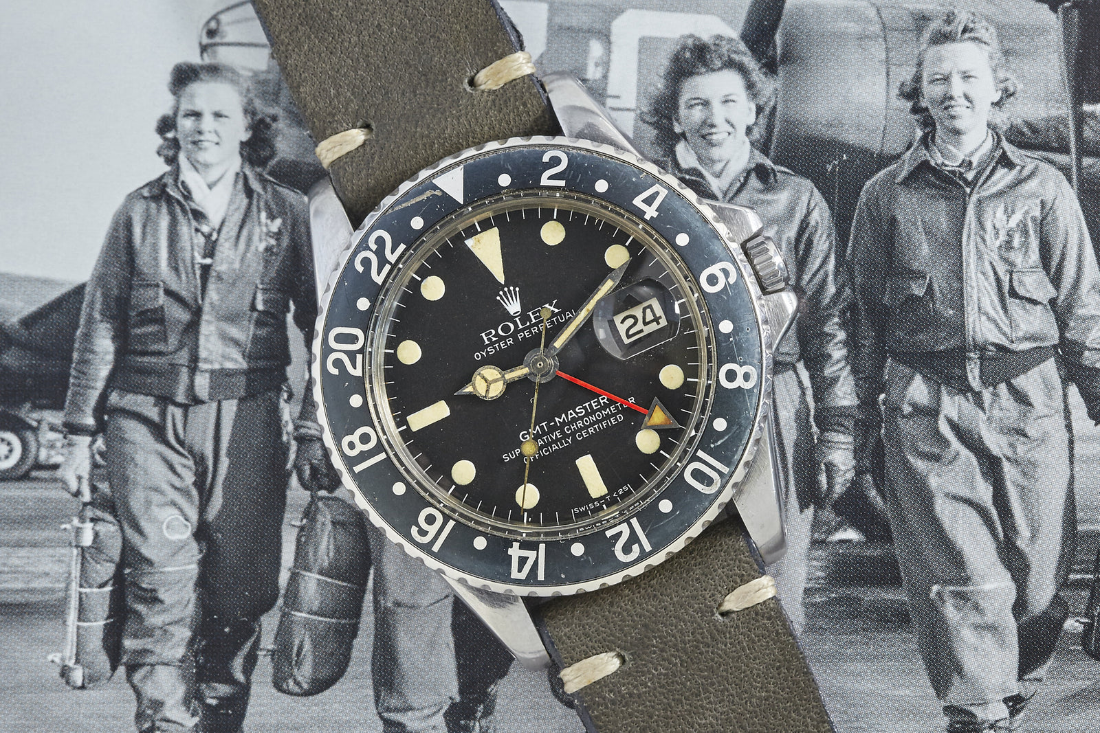 A Rolex GMT-Master by Rolex with a black bezel and leather strap is displayed atop a black-and-white photo of three smiling female pilots in flight suits, standing in front of an aircraft.