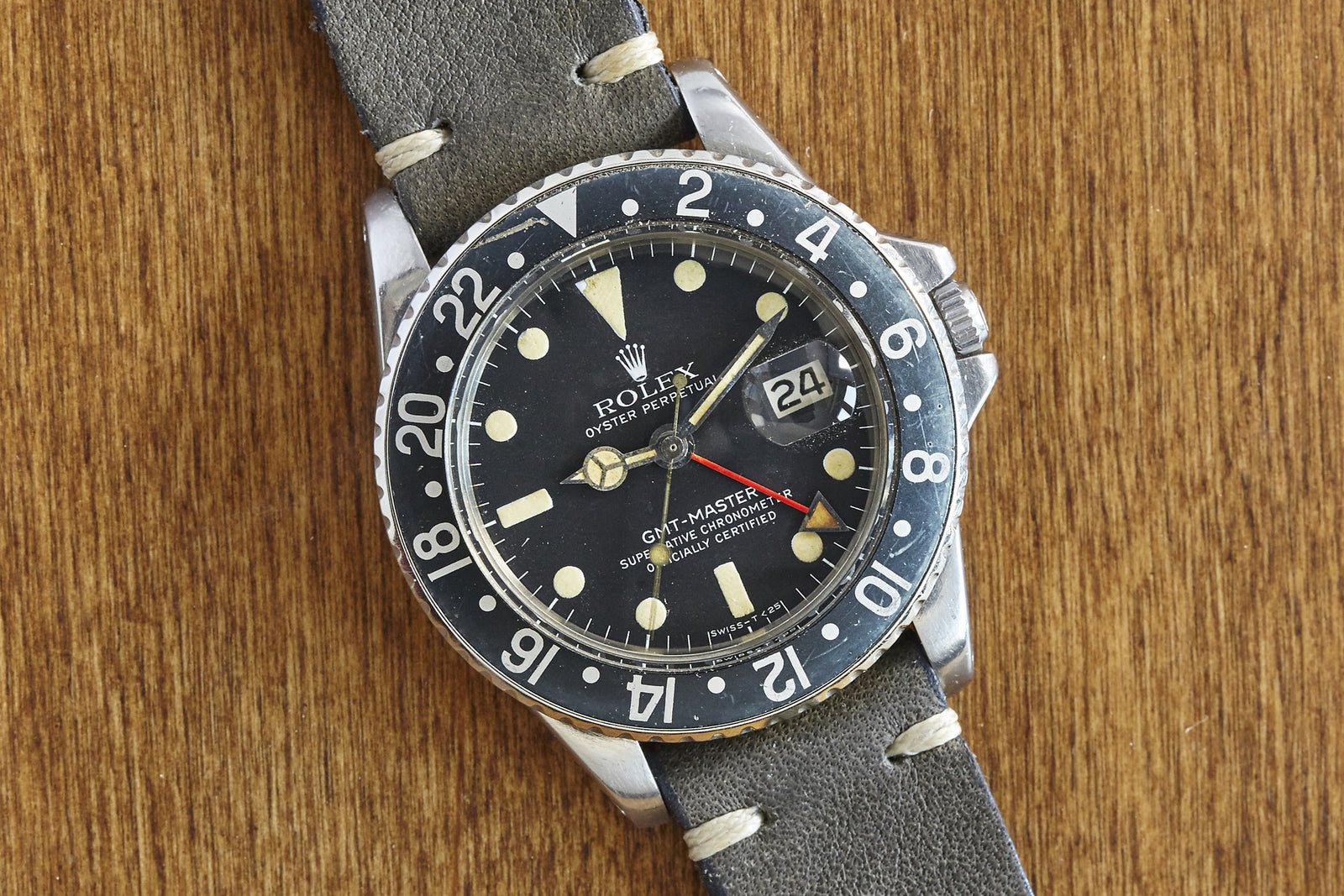 The Rolex GMT-Master by Rolex features a black dial, red and white hands, and a black bezel on a grey leather strap, shown on a wooden surface with a magnified date display—ideal for vintage Porsche fans.