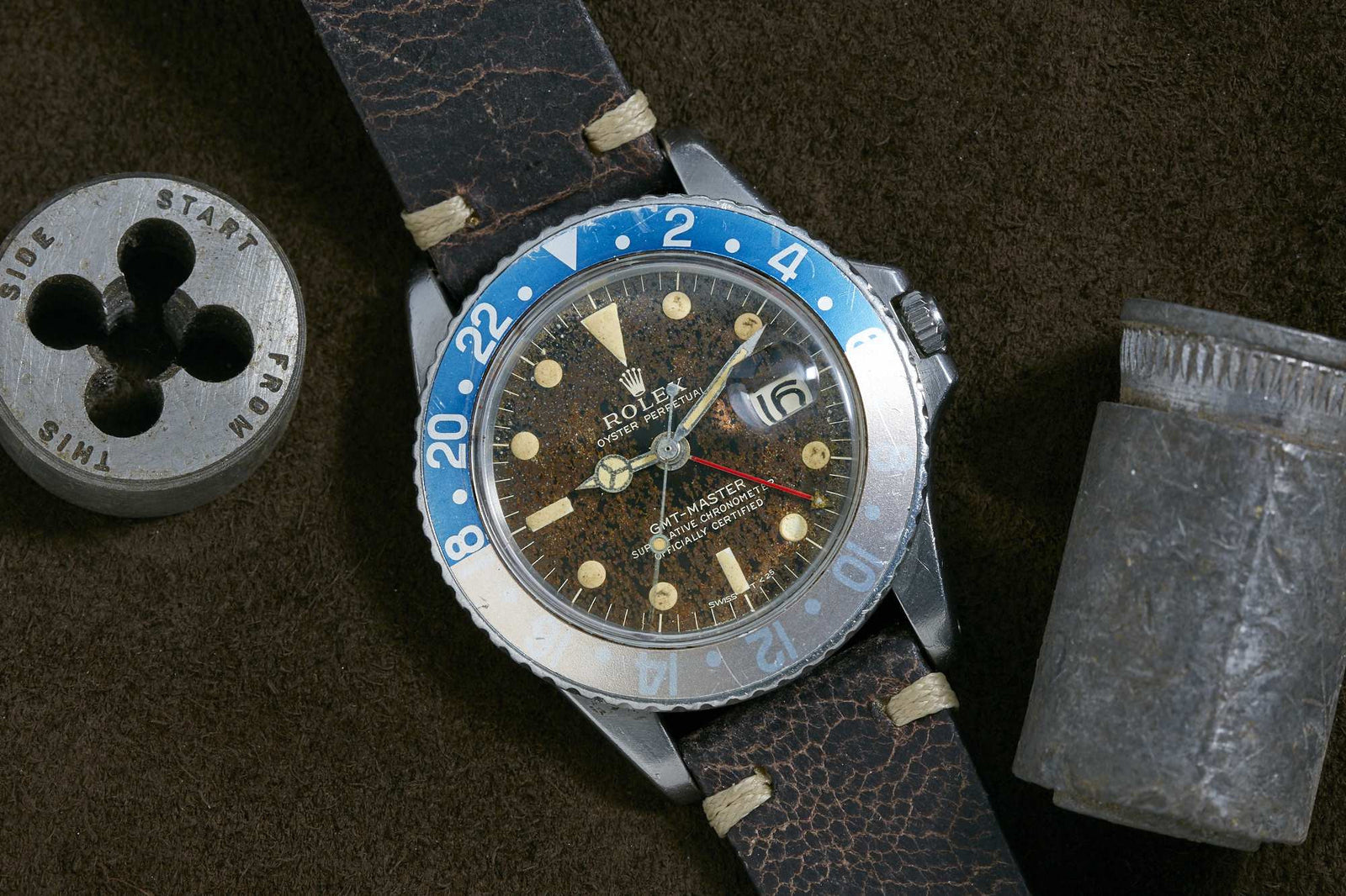 A Rolex GMT-Master wristwatch with faded blue and white bezel, aged brown dial, and leather strap sits on a brown surface next to a metal tool, highlighting the classic style of this iconic Rolex sport watch.