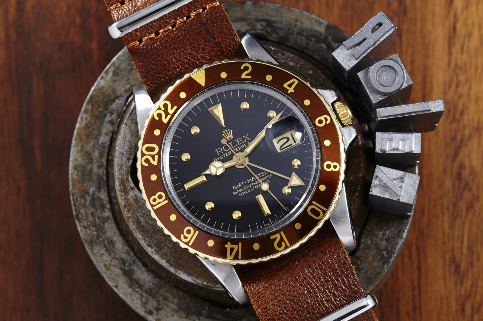 A vintage Rolex GMT Master 1675 "Root Beer" Nipple Dial with a brown leather strap and brown-gold bezel is displayed on a metal surface, with metal letter stamps spelling ROLEX beside it.