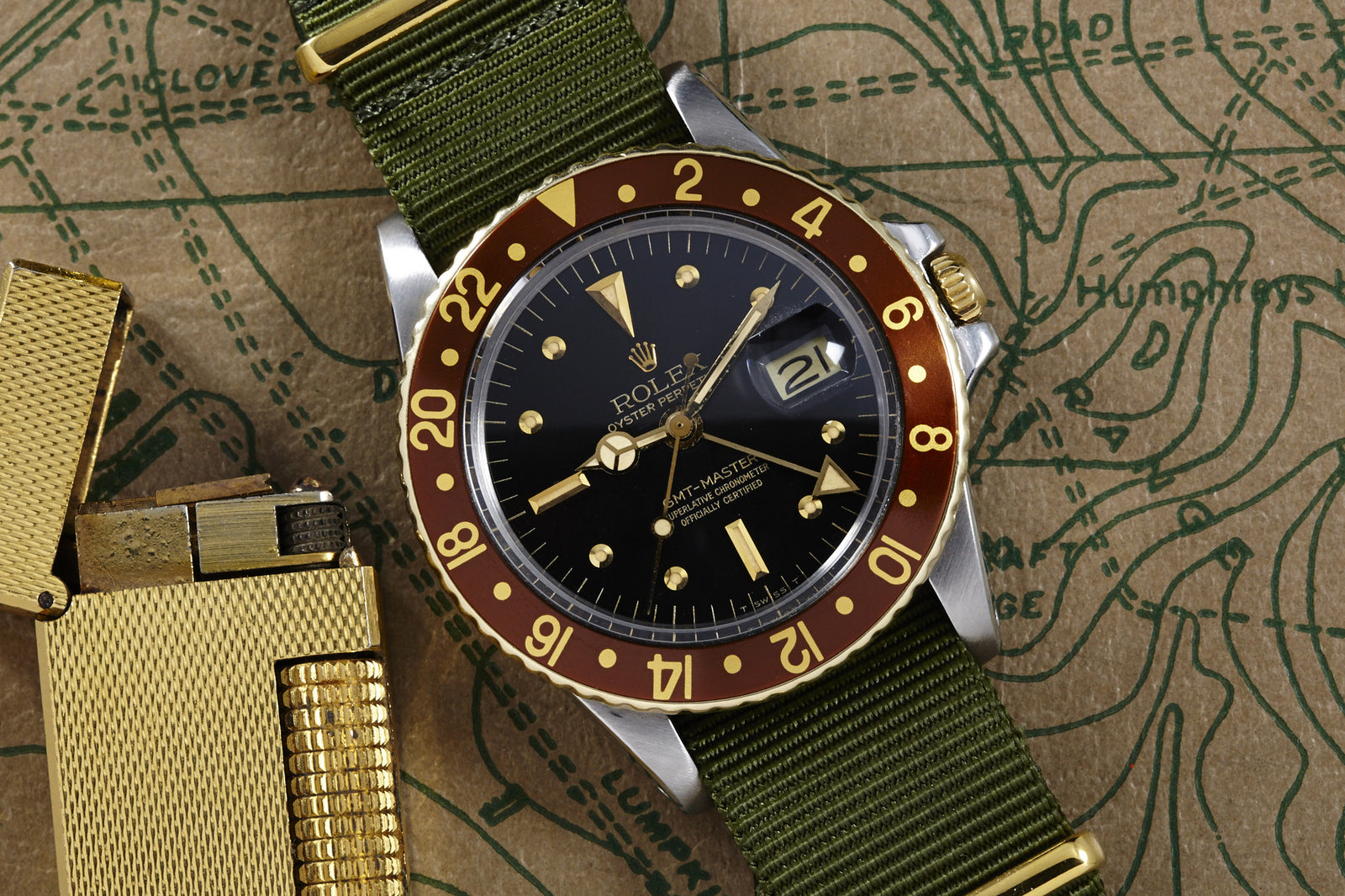 A Rolex GMT Master 1675 "Root Beer" Nipple Dial with a red and gold bezel, black dial, and green fabric strap rests on a vintage map beside a gold lighter. This two-tone Rolex displays the date and features gold hour markers.