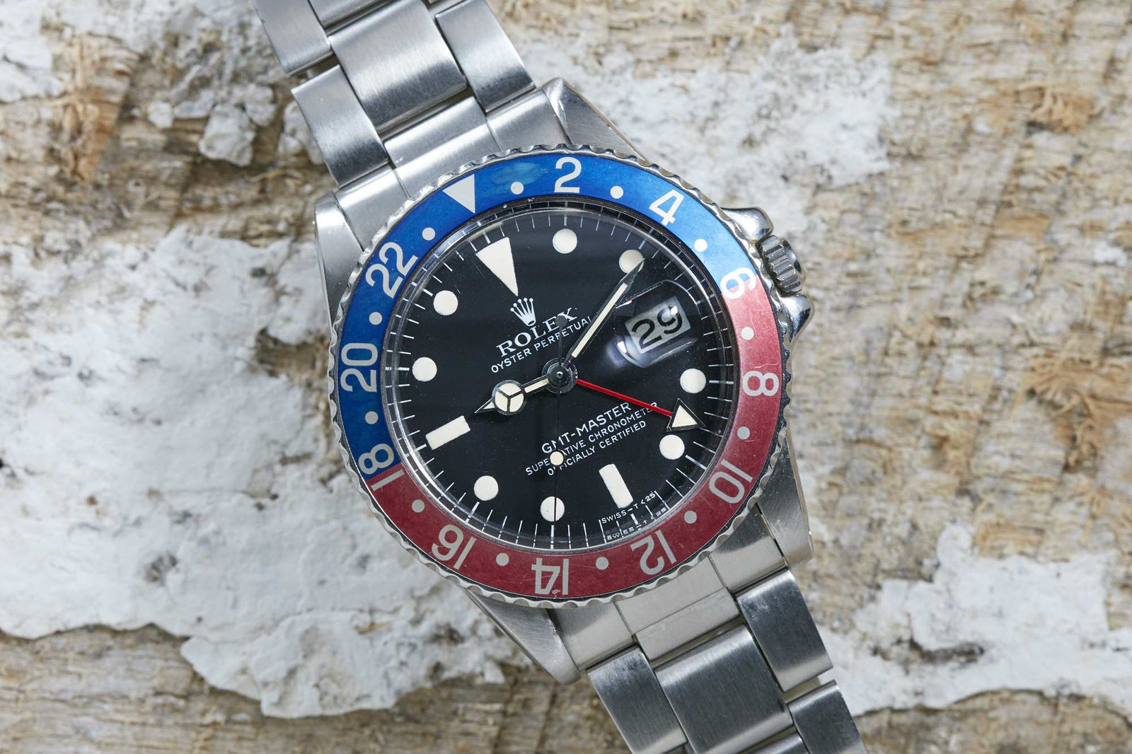 A Rolex GMT Master by Rolex with a red and blue bezel, black dial, and date display sits on a weathered wooden surface with white patches.
