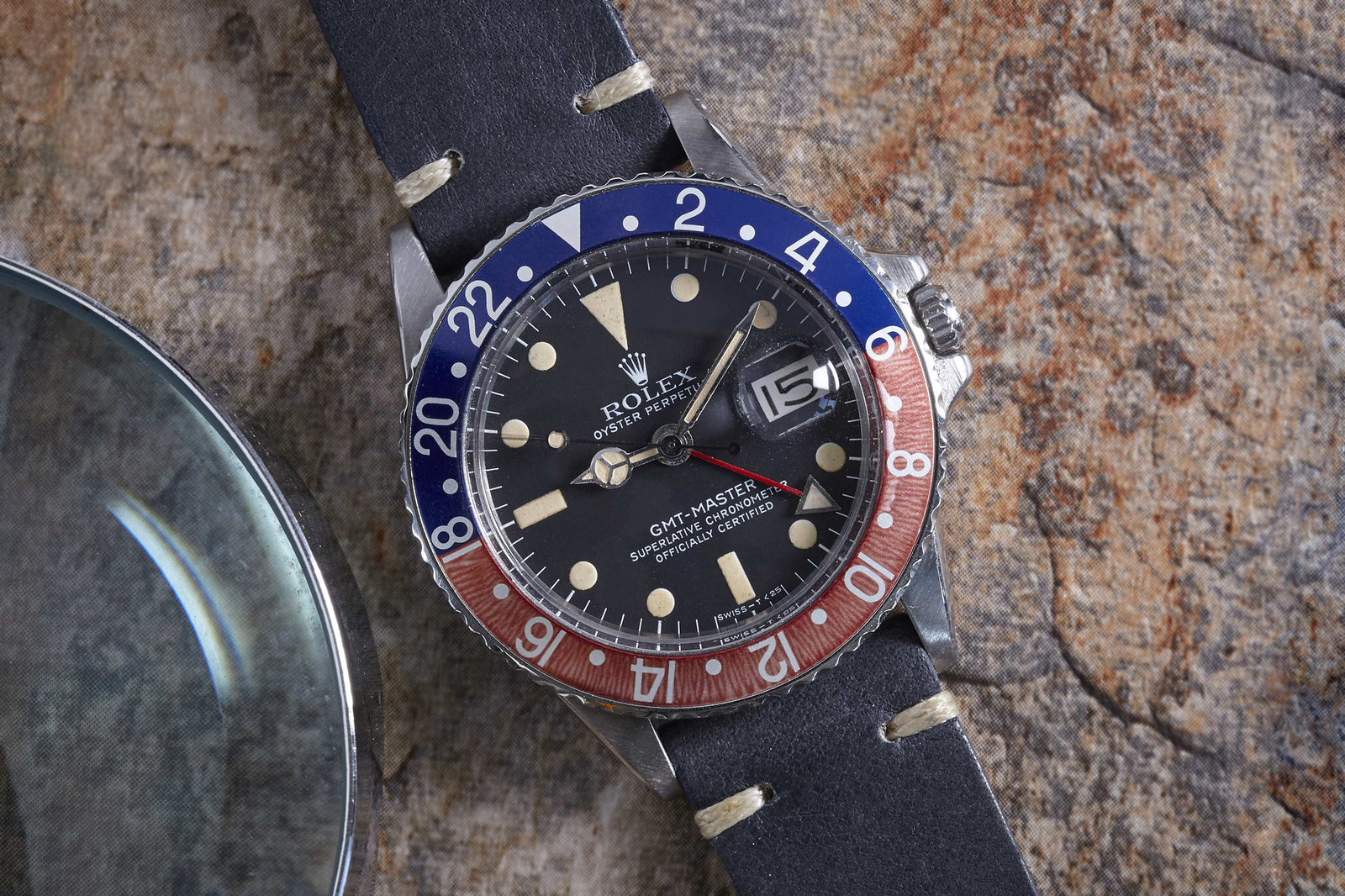 The Rolex GMT-Master by Rolex, a classic among vintage and pilot watches, features a black dial, red and blue bezel, leather strap, and signature date window. It sits on textured stone next to a magnifying glass.