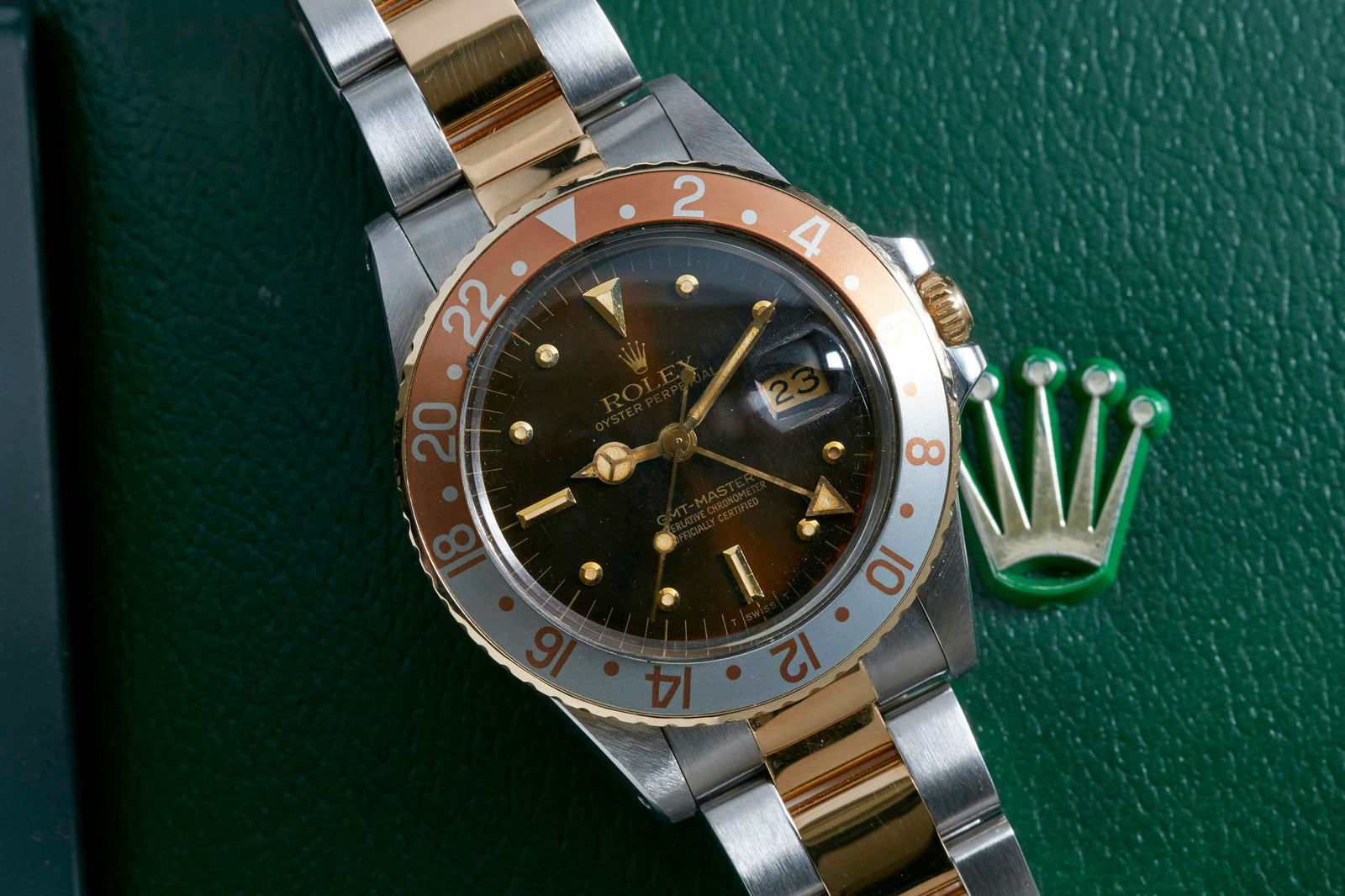 The Rolex GMT Master Ref. 16753 Box and Papers is a two-tone sports watch with a stainless steel case, brown and gold bezel, brown dial, gold hour markers, displayed on green beside the signature Rolex crown logo.