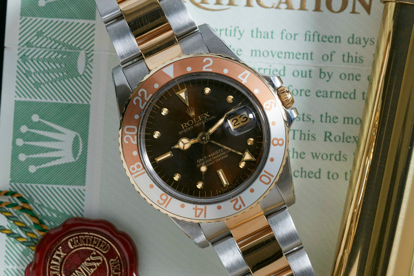 The Rolex GMT Master Ref. 16753, featuring a black and gold dial, is displayed with its box, papers, and a red wax seal beside its elegant stainless steel case. The date on the watch face shows 23.