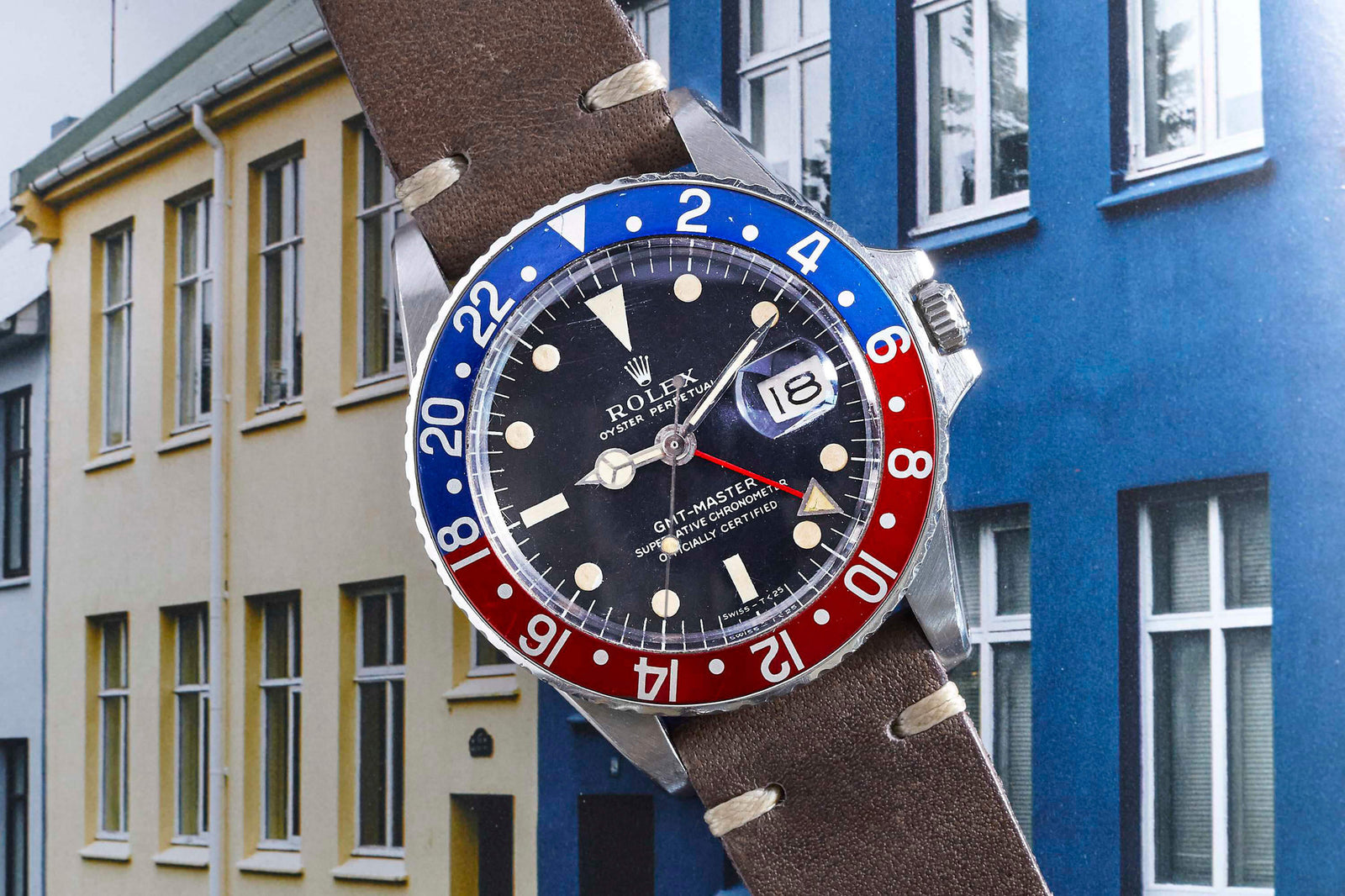 A Rolex GMT-Master by Rolex, featuring a brown leather strap and a red and blue bezel, rests atop a book with yellow and blue buildings with many windows.