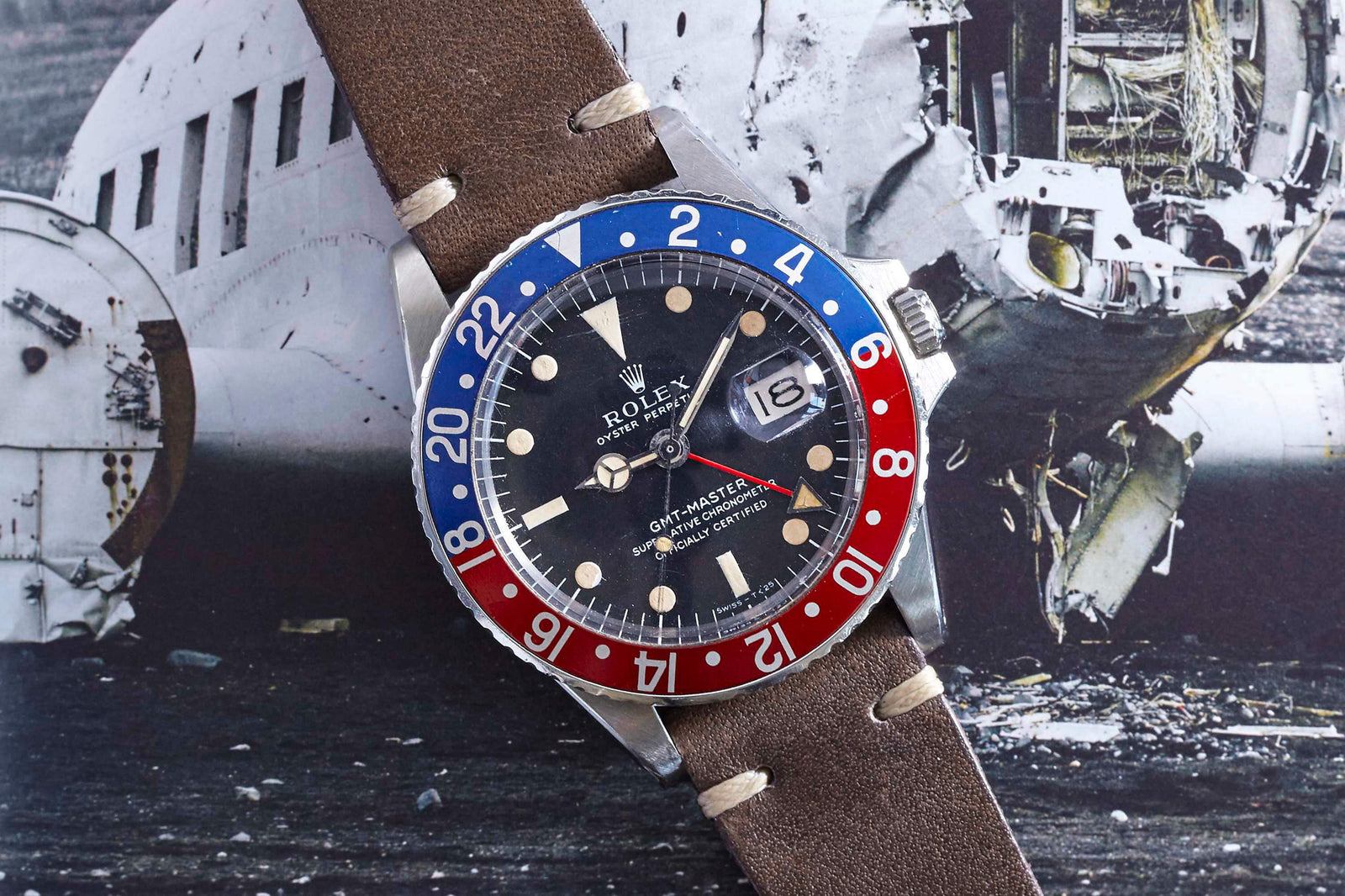 A Rolex GMT-Master by Rolex featuring a red and blue bezel with a brown leather strap is displayed atop a black-and-white photograph of a damaged airplane.