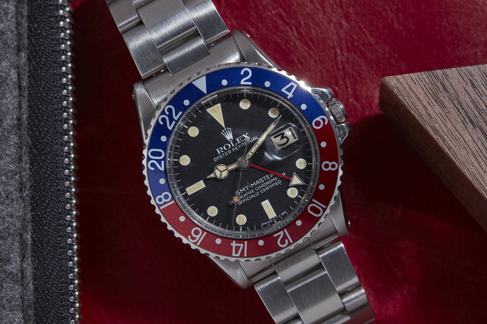 A close-up of the Rolex GMT Master by Rolex, featuring a red and blue bezel, black dial, and silver bracelet, displayed on a red surface beside a wooden object. The watch date reads 31.