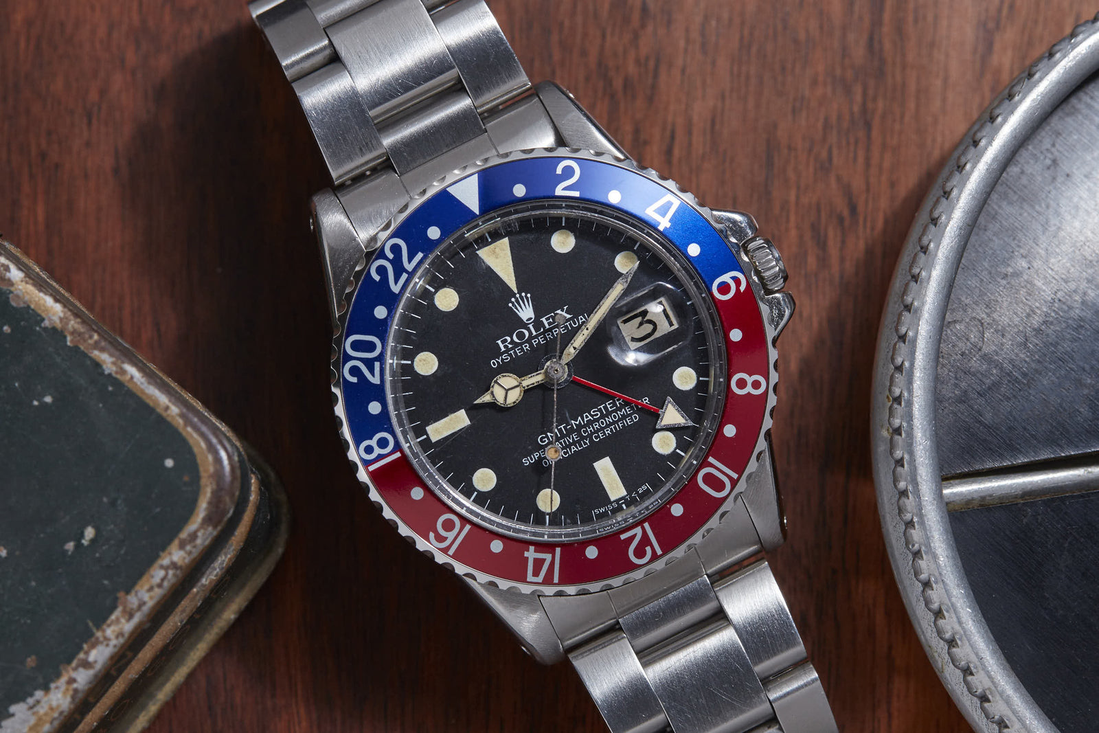 A Rolex GMT Master by Rolex, featuring a stainless steel band and a red-and-blue bezel, is displayed on a wooden surface next to a metal item and the edge of a green box.