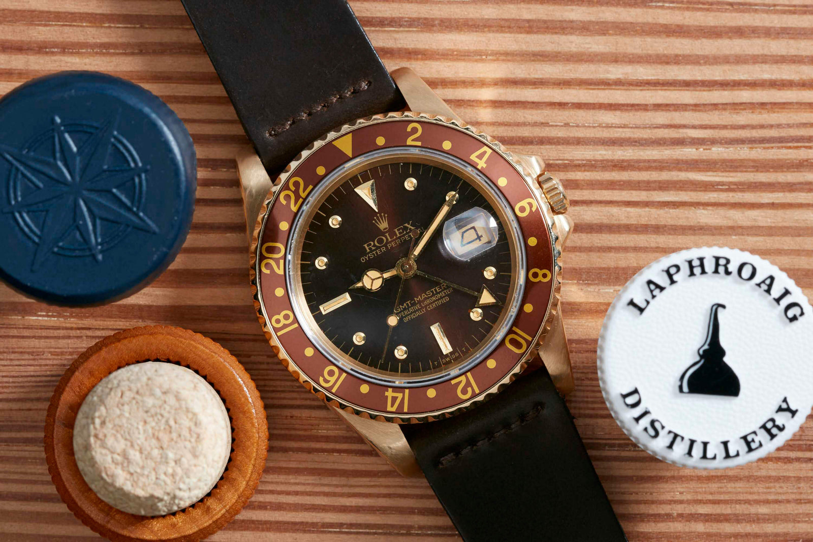 A Rolex GMT Master 18K wristwatch with a black and red bezel, Sport Oyster case, and brown leather strap rests on wood beside a blue wax seal, cork, and a white Laphroaig Distillery bottle cap.
