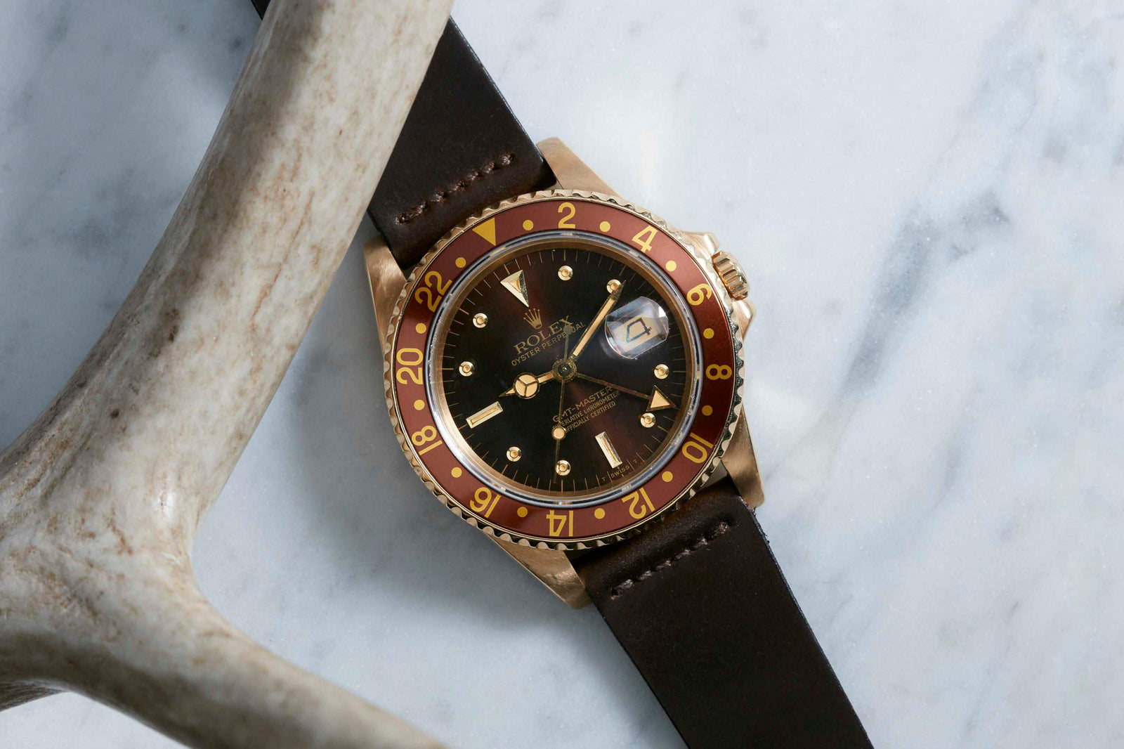 A Rolex GMT Master 18K wristwatch with a brown and gold bezel and dark leather strap, featuring the iconic Oyster case, rests on a white marble surface beside a light-colored antler.