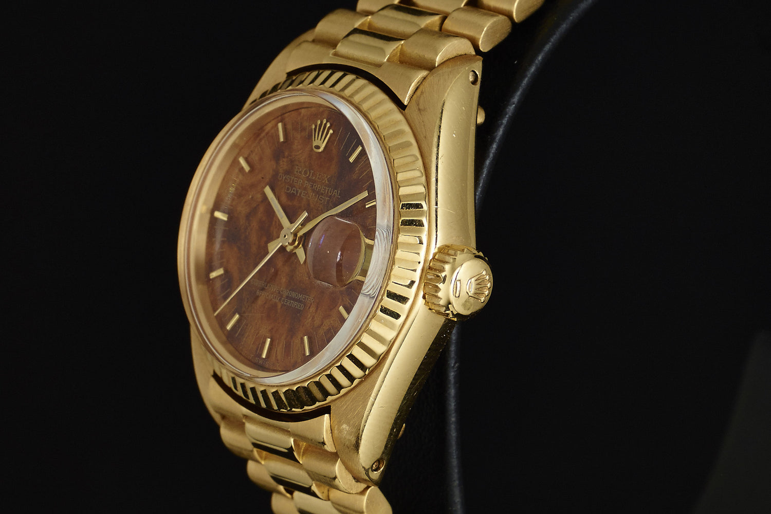 A Rolex Lady Datejust 18K Mahogany Dial wristwatch with gold markers, fluted bezel, and gold bracelet is shown at an angle against a dark background.