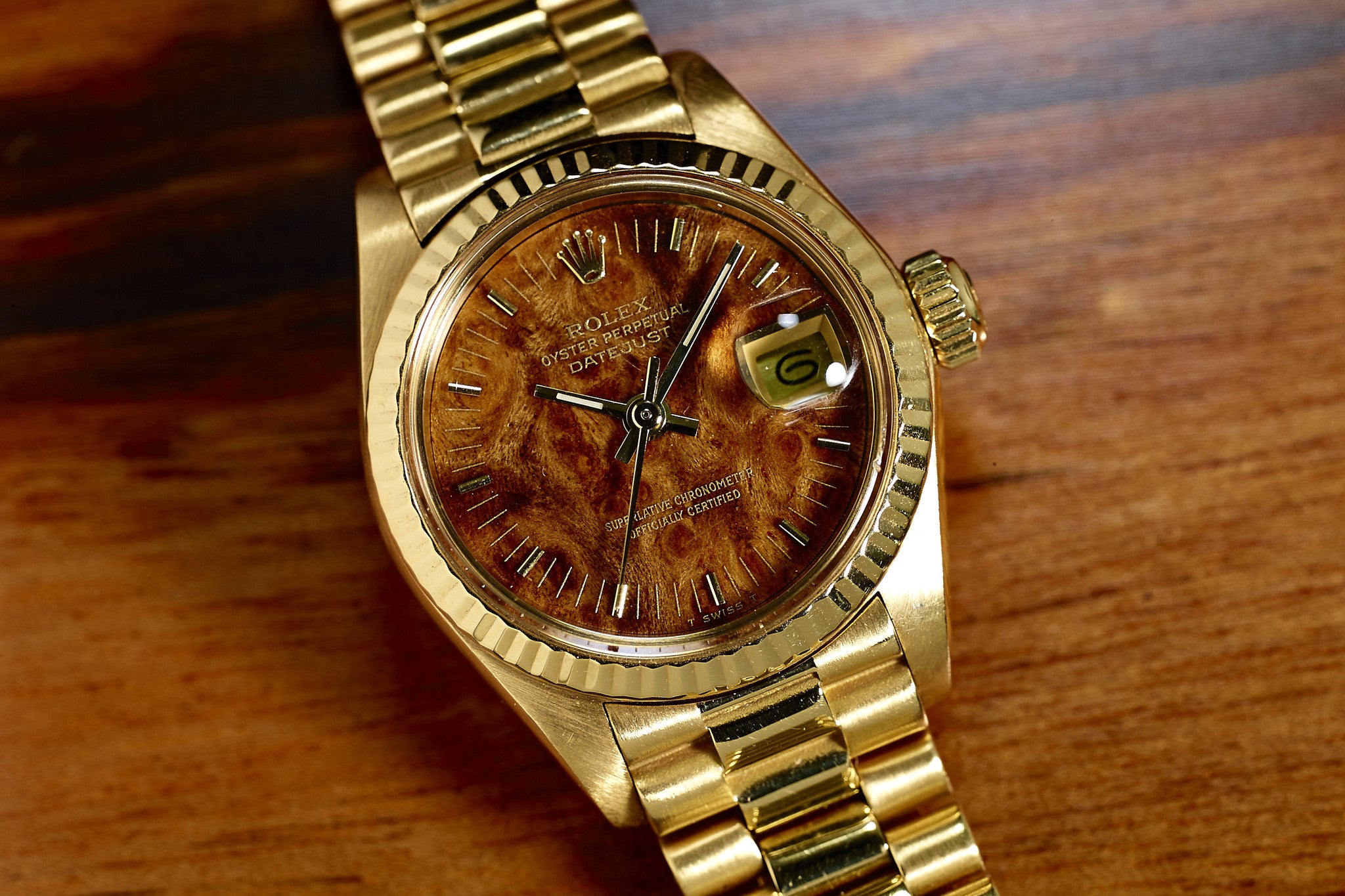 The Rolex Lady Datejust 18K Mahogany Dial features a unique brown wood-patterned dial, gold metal band, and date window showing 6. This vintage ladies’ watch by Rolex is displayed on a wooden surface.