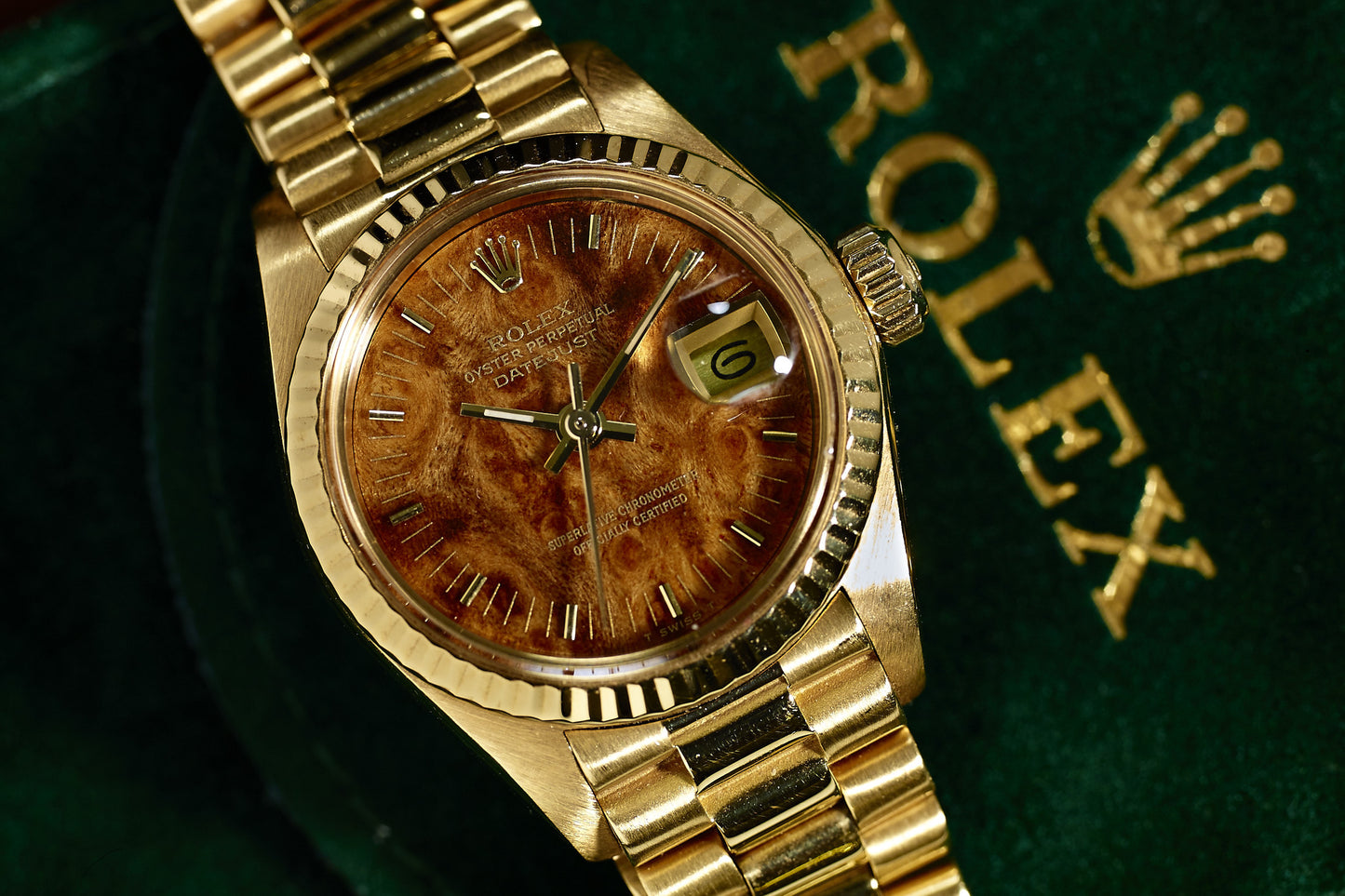 The Rolex Lady Datejust 18K Mahogany Dial features a textured brown dial and date display, elegantly displayed on a green surface with the gold Rolex logo and crown.