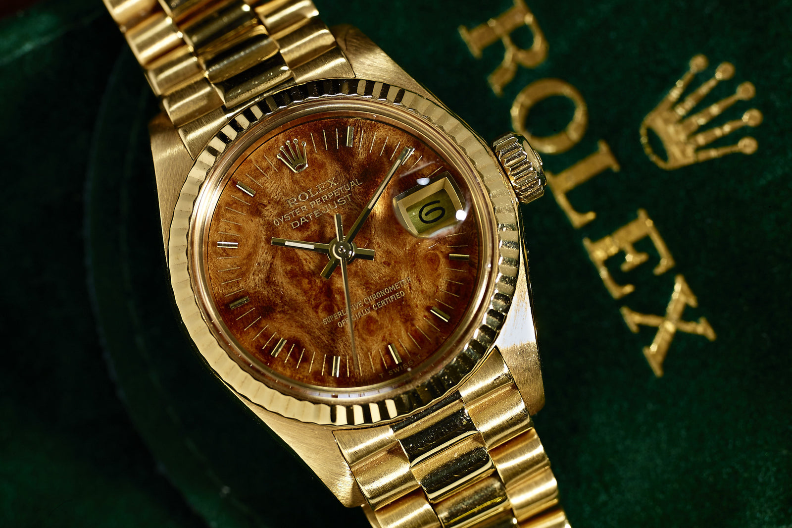 The Rolex Lady Datejust 18K Mahogany Dial features a textured brown dial and date display, elegantly displayed on a green surface with the gold Rolex logo and crown.