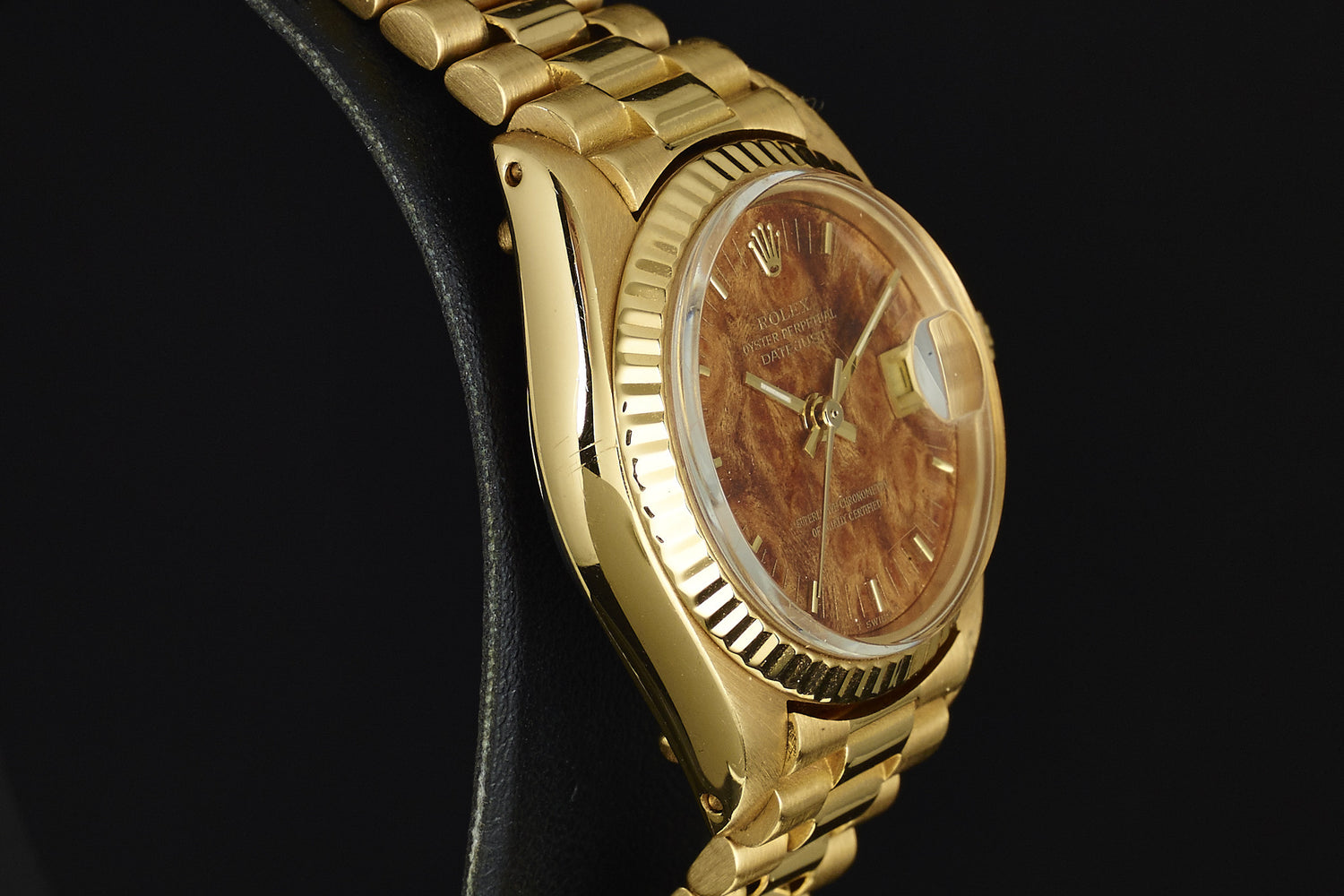 A close-up of the Rolex Lady Datejust 18K Mahogony Dial wristwatch features a gold bracelet, brown mahogany-textured dial, and fluted bezel, elegantly displayed on a black stand against a dark background.