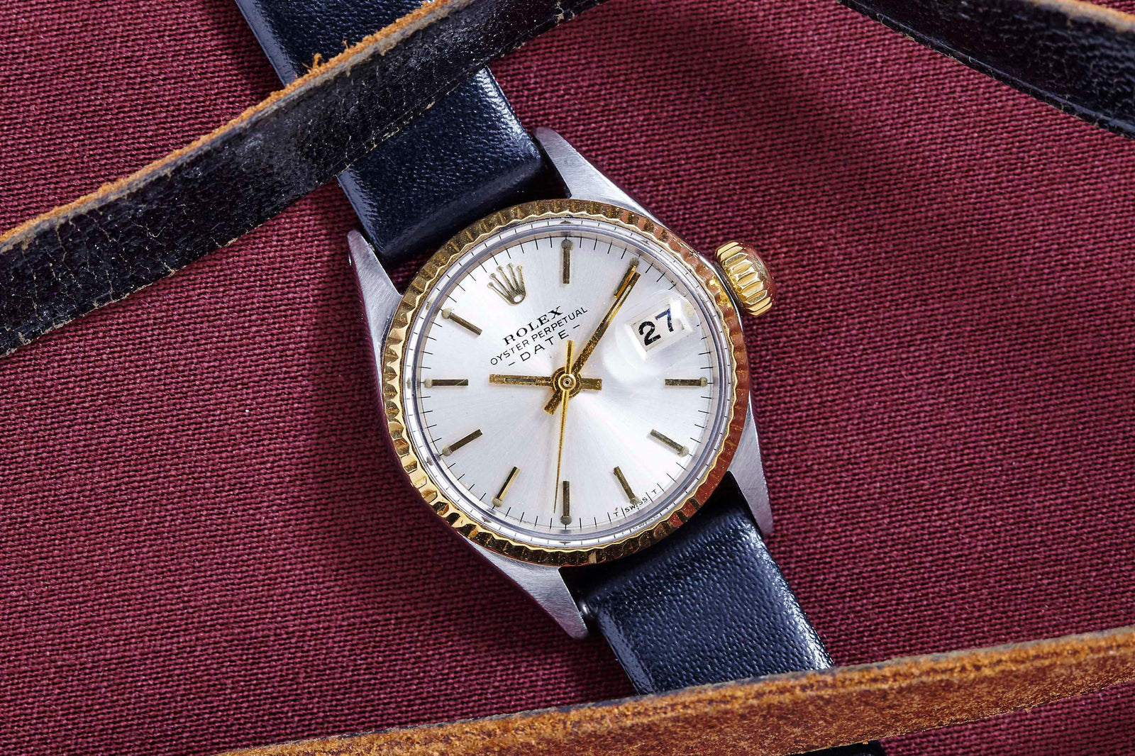 A Rolex Lady Oyster Perpetual Date women's watch with a silver dial, gold bezel, and black leather strap is displayed on burgundy fabric, partially covered by two brown leather straps—a timeless vintage piece from Rolex.