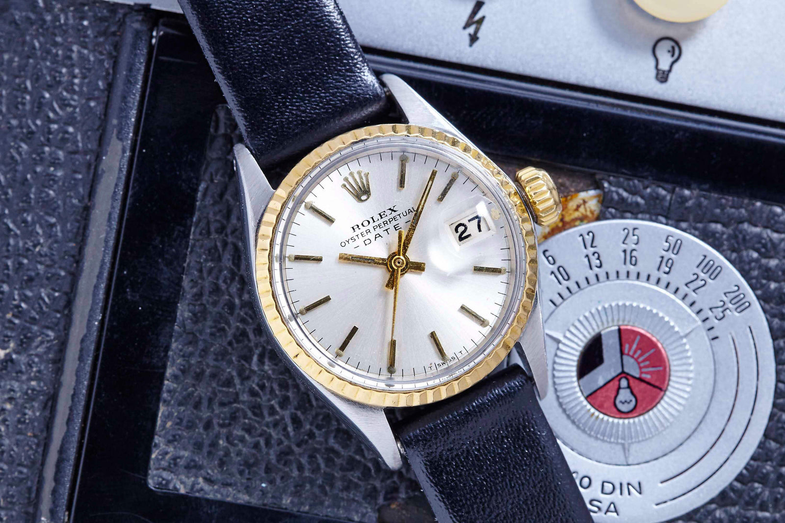 A close-up of a Rolex Lady Oyster Perpetual Date watch with a silver dial, gold bezel, black leather strap, and date at 3 o’clock, shown resting on a vintage camera with a visible temperature gauge.