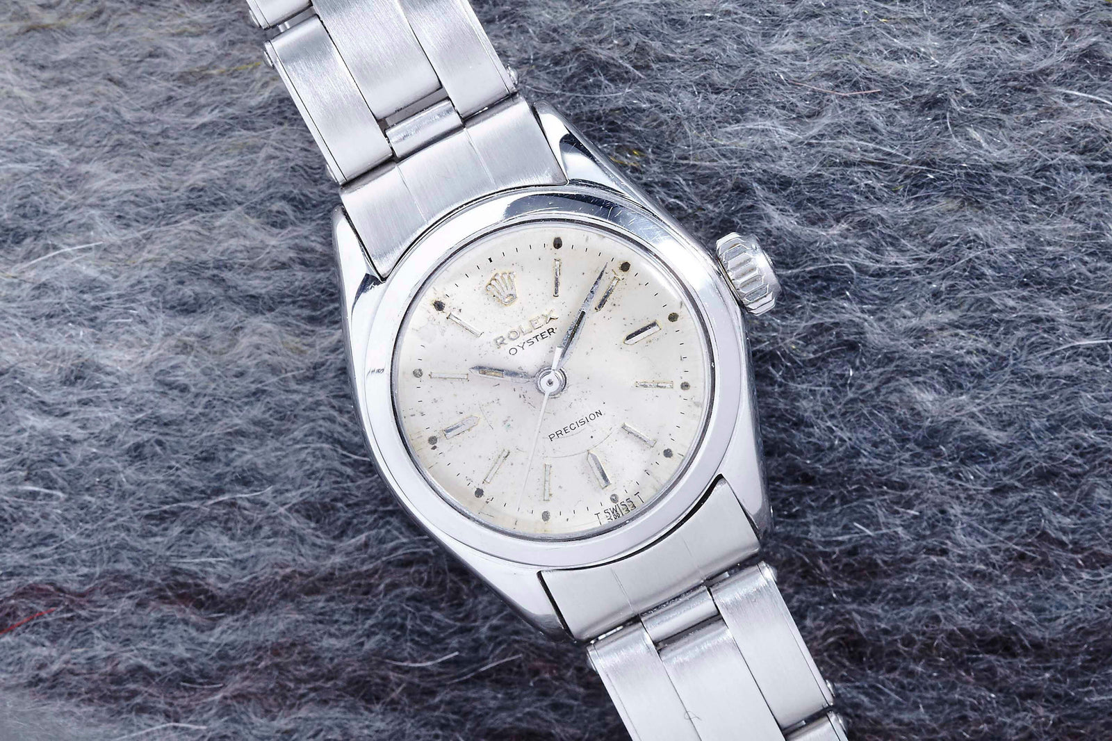 The Rolex Lady Oyster Precision by Rolex features a silver metal band and white dial with vintage patina, displayed on a textured gray surface.