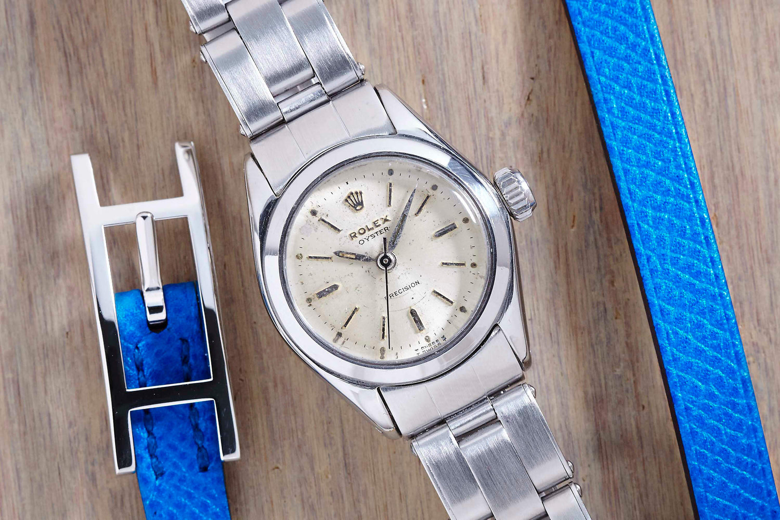 A Rolex Lady Oyster Precision wristwatch by Rolex with a metal bracelet rests on a wooden surface beside a blue leather strap featuring a silver clasp.
