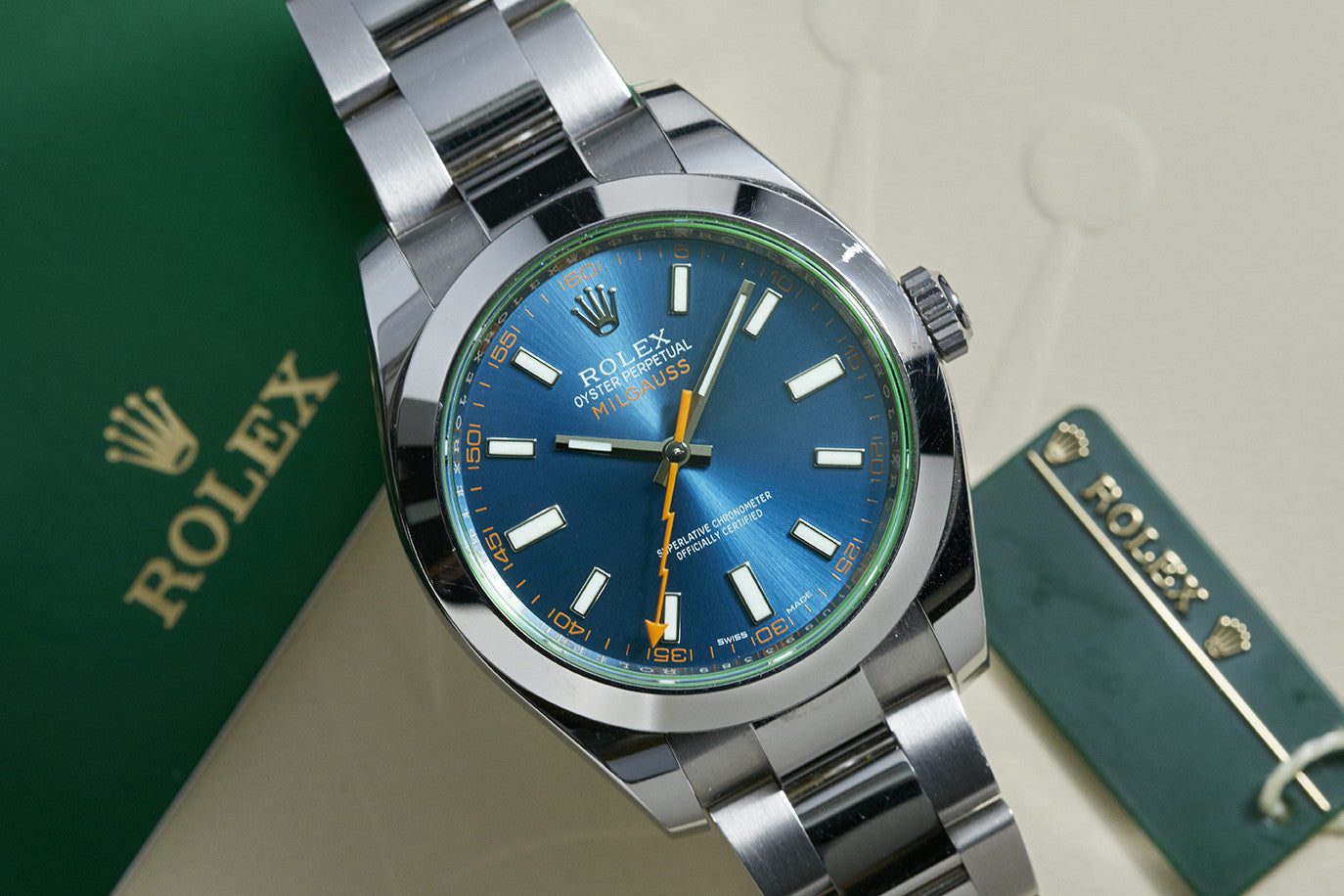 A close-up of the Rolex Milgauss by Rolex, featuring a blue dial, green-tinted sapphire crystal, and orange lightning-bolt second hand, displayed next to a green Rolex booklet and tag.
