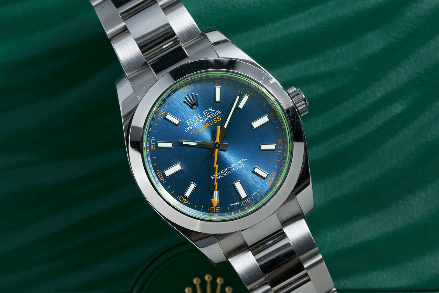 A close-up of the Rolex Milgauss by Rolex, featuring a metallic band, blue dial, orange lightning bolt seconds hand, and white hour markers, displayed on a green textured background.