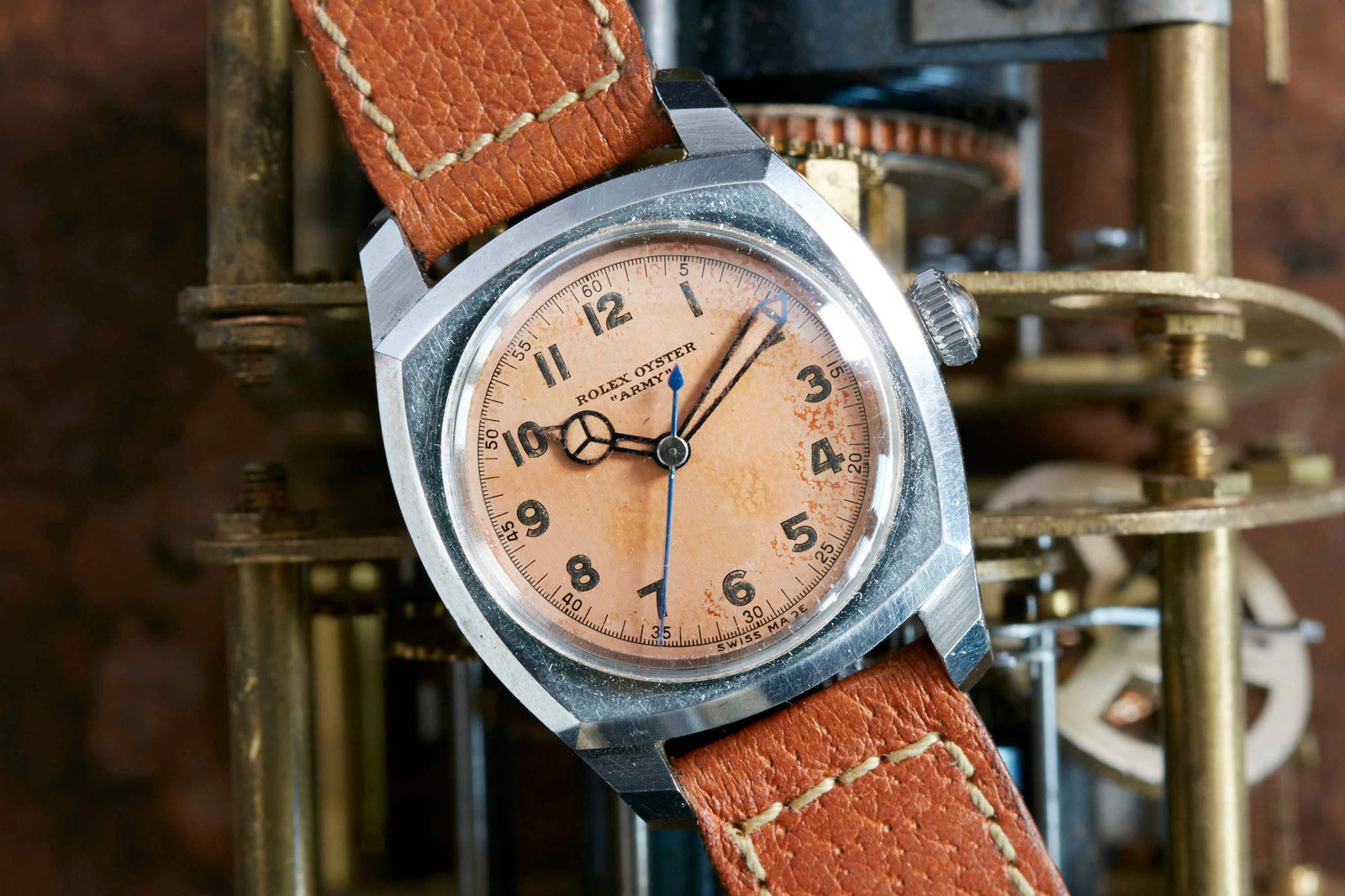 The Rolex Oyster Army by Rolex features a weathered pink dial and brown leather strap, showcased against intricate brass mechanics, evoking the look of classic World War II military watches.