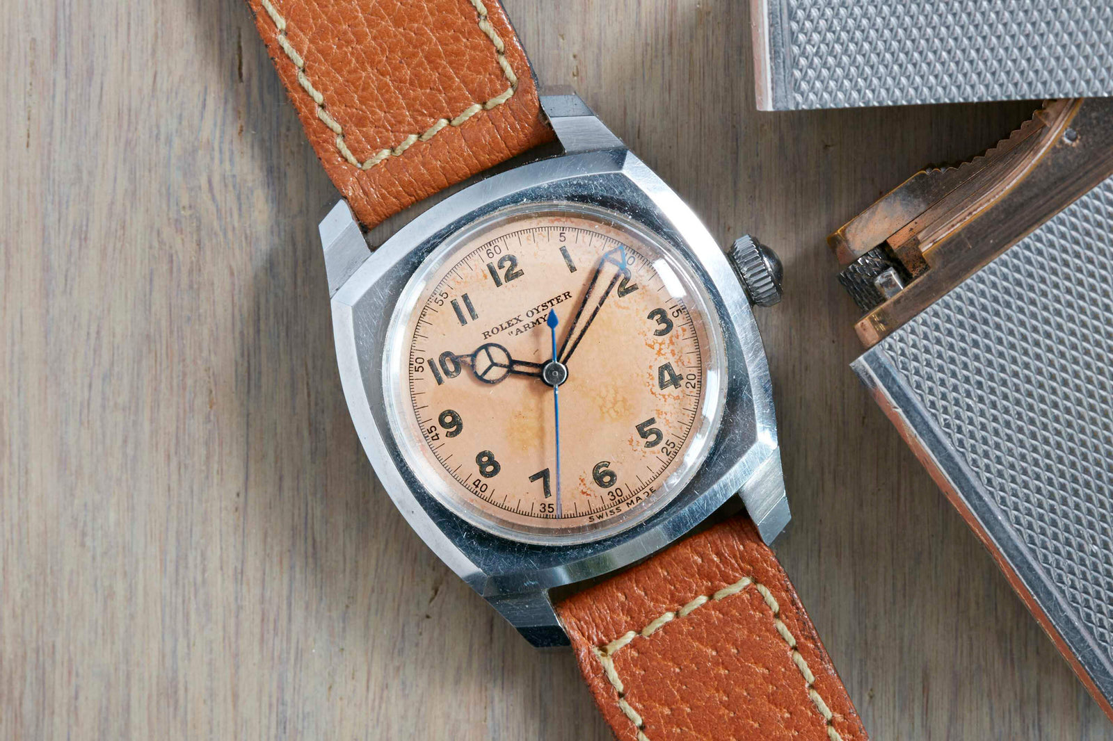 A vintage Rolex Oyster Army watch with a worn light brown dial, black numerals, and a tan leather strap rests on wood beside a textured metal object, echoing the classic World War II style of Rolex military timepieces.
