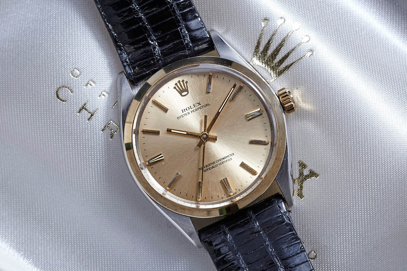 A vintage Rolex Oyster Perpetual wristwatch featuring a gold dial, gold hour markers, and a black leather strap is displayed on a satin Rolex pouch embossed with the brand’s crown logo.