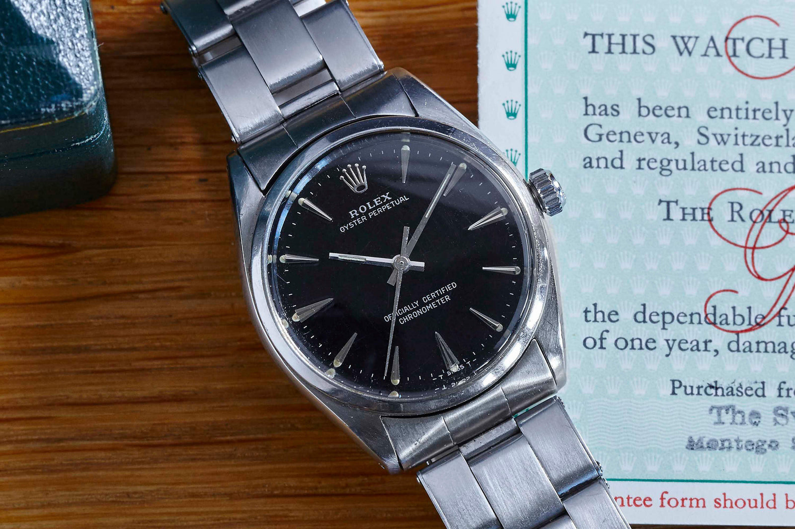 A Rolex Oyster Perpetual by Rolex featuring a stainless steel case and black dial is placed on a wooden surface next to an official document with blue patterned borders.