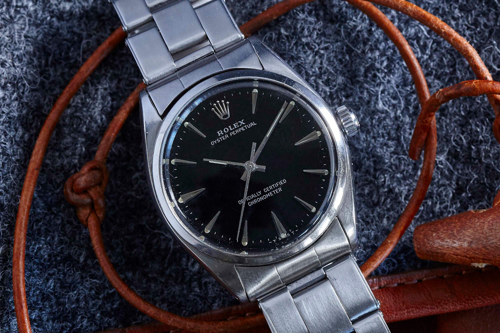 A Rolex Oyster Perpetual wristwatch by Rolex, featuring a stainless steel case, black dial, and silver hour markers, rests on textured gray fabric with brown leather straps partly visible in the background.