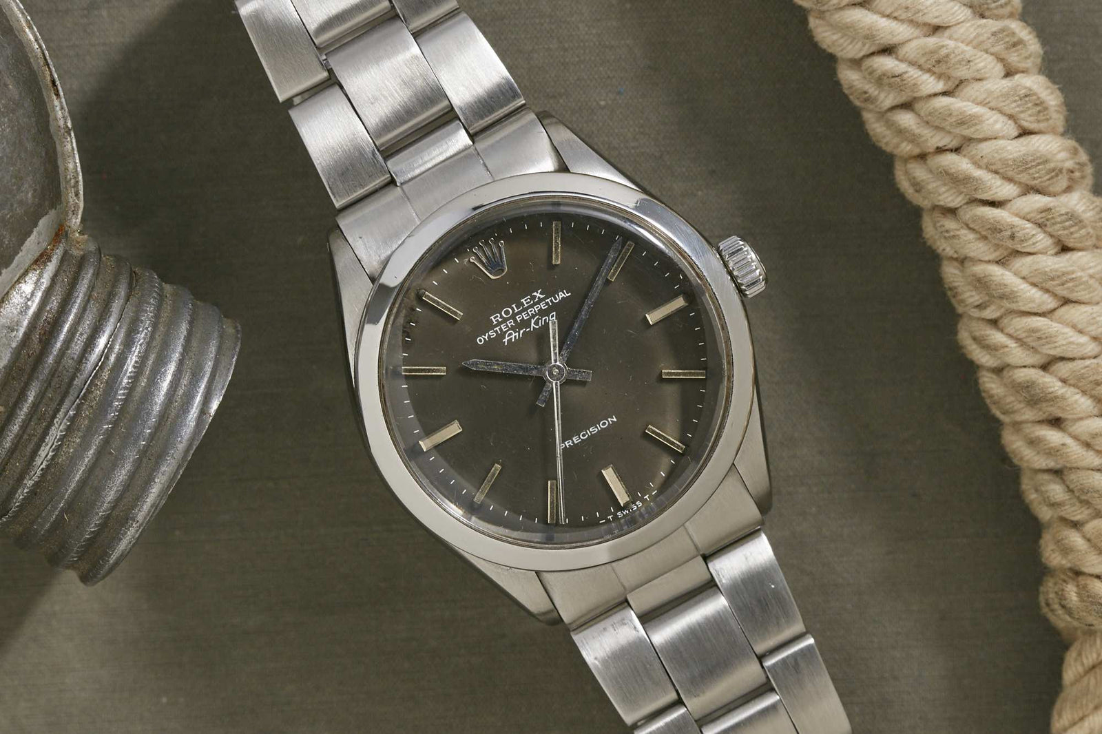 A Rolex Oyster Perpetual Air King wristwatch with a stainless steel band and dark dial is displayed on a gray surface beside a coiled rope and metallic object, showcasing the timeless appeal of the Rolex Oyster Case.