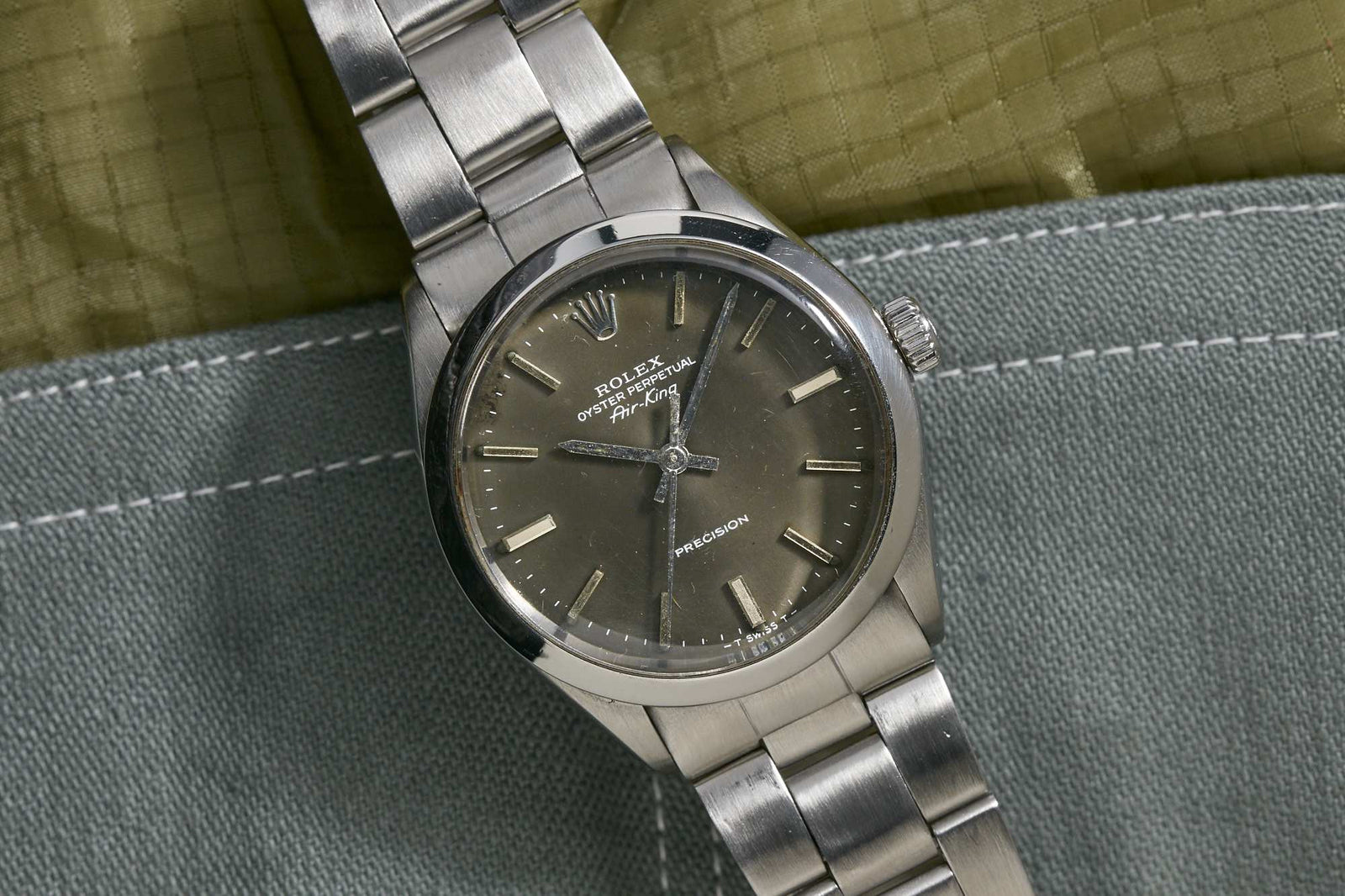 A Rolex Oyster Perpetual Air King wristwatch with a dark gray dial, silver hour markers, and a metal bracelet rests on green and gray fabric, highlighting the timeless style of the classic Rolex Oyster Case.