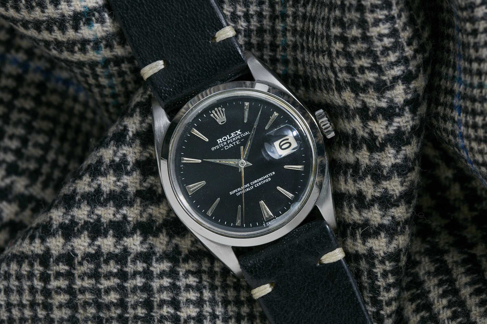 A Rolex Oyster Perpetual Date watch with a black dial, perpetual movement, and black leather strap by Rolex lies on black-and-white houndstooth fabric. The magnified date is at 3 o’clock.