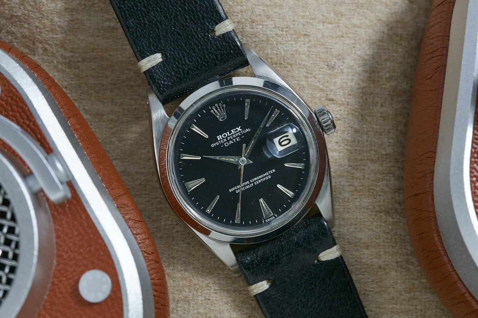 A Rolex Oyster Perpetual Date by Rolex with a black dial and leather strap rests on a tan surface, positioned between brown and silver headphones.