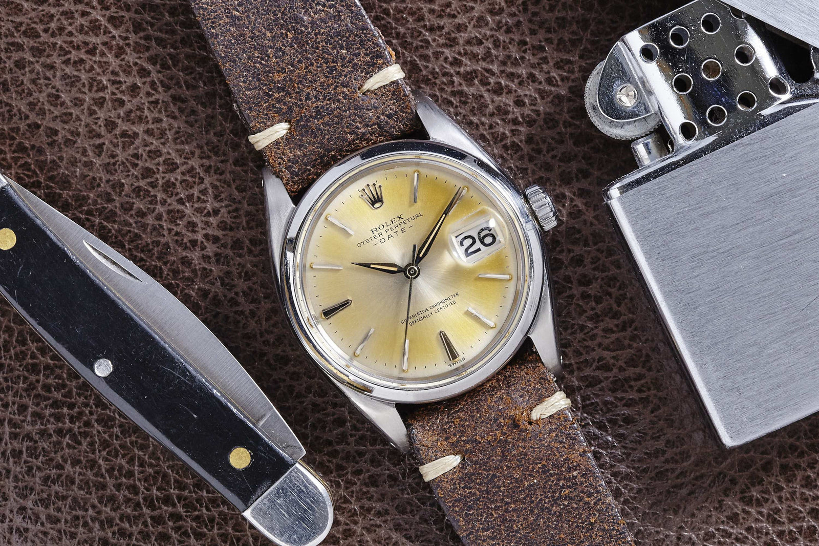 A Rolex Oyster Perpetual Date by Rolex, featuring a silver sunburst dial and worn brown leather strap, rests on textured brown leather beside a folding pocket knife and silver metal lighter for a classic touch.