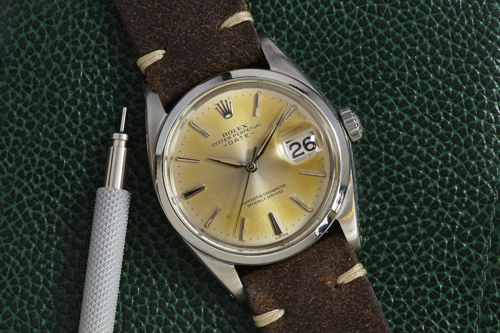 A Rolex Oyster Perpetual Date by Rolex, featuring a silver sunburst dial, brown leather strap, and date display, rests on a textured green surface next to a small metal tool.