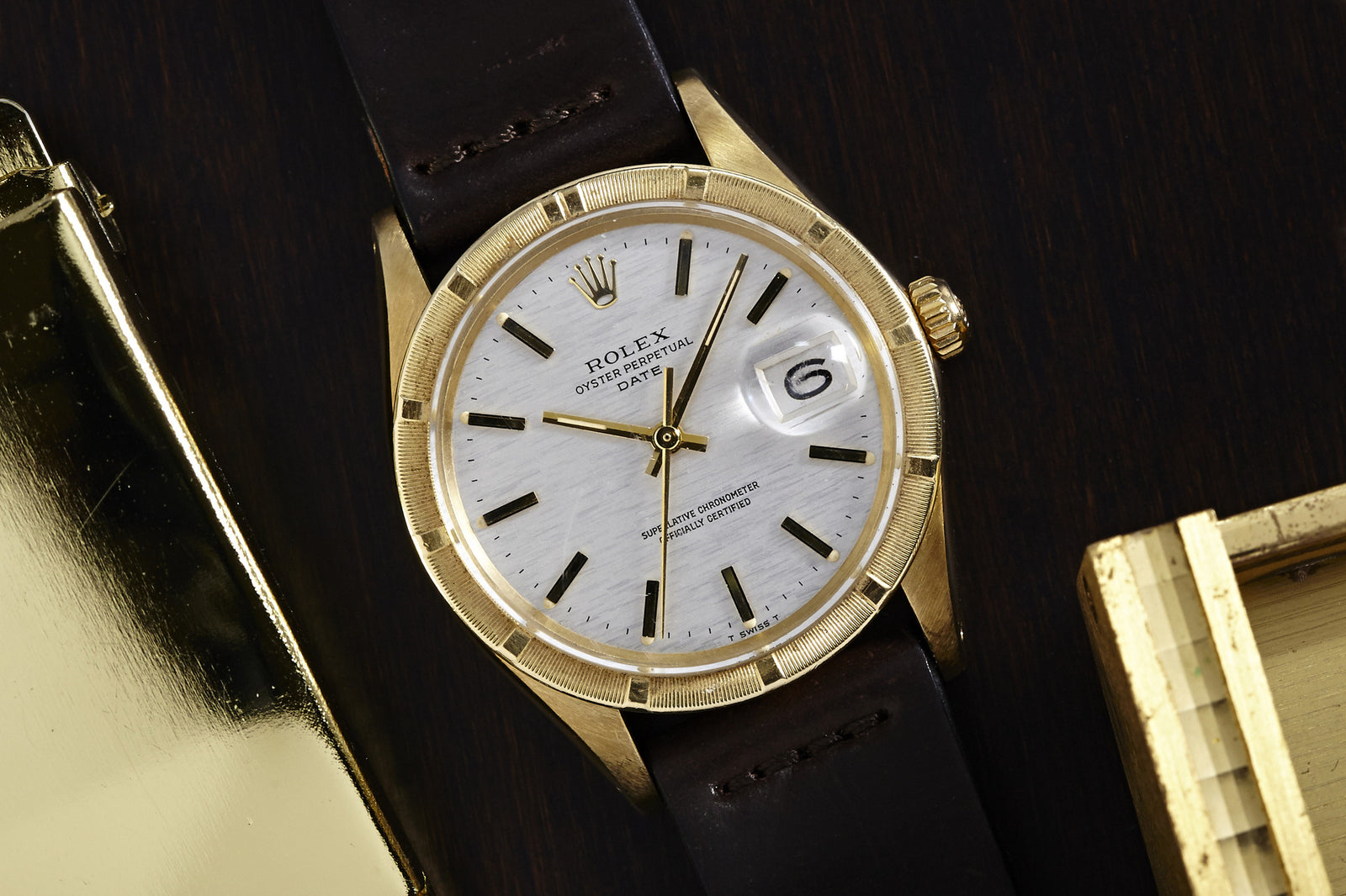 A Rolex Oyster Perpetual Date Gold Linen Dial Ref. 1501 watch by Rolex, featuring a gold linen-textured dial and black leather strap, sits on a dark wooden surface near two gold objects.