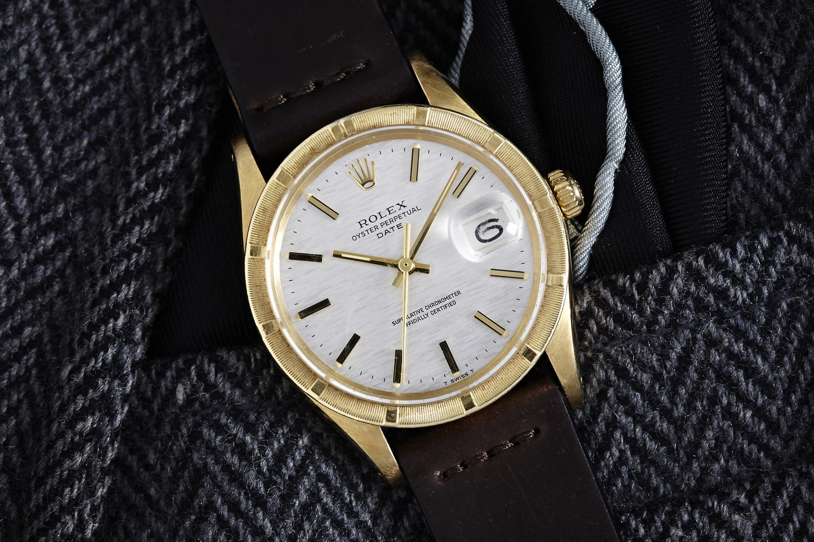 A Rolex Oyster Perpetual Date Gold Linen Dial Ref. 1501 watch with a gold case and brown leather strap is shown on gray herringbone fabric, featuring the date at 3 o’clock.
