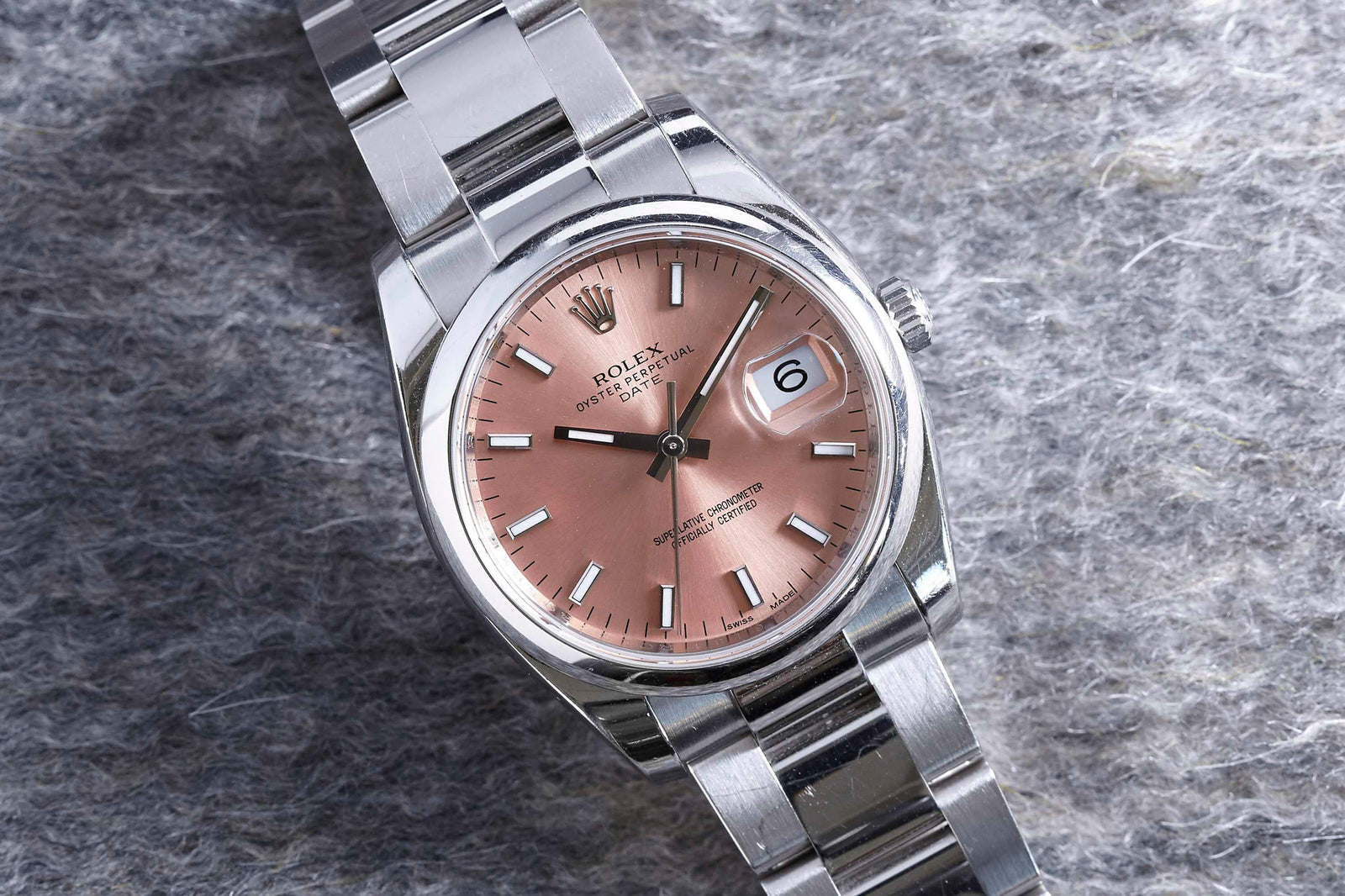 The Rolex Oyster Perpetual Date, a luxury women’s watch by Rolex, features a pink dial and date display. Shown on a textured gray surface, this stainless steel collectible displays the time as approximately 2:08.