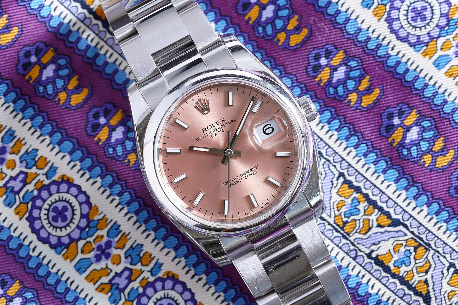 A Rolex Oyster Perpetual Date, a luxury ladies watch from Rolex, with a pink dial and silver markers, rests on vibrant purple and yellow patterned fabric.